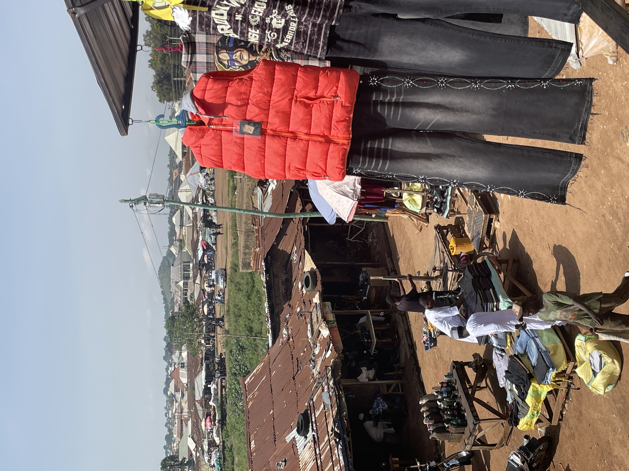 The image captures a bustling open-air market in a city, likely in a developing country, given the rudimentary structures and the general atmosphere. In the foreground, a stall displays various clothing items, including a bright red puffer vest and several pairs of dark denim jeans embellished with decorative stitching and rhinestones. The jeans are hung on a rack, with their frayed hems visible. Behind this stall, the market unfolds with more stalls covered by corrugated metal roofs and tarps, selling a variety of goods. There are piles of folded clothing, barrels of liquids, and a display of what appear to be jerrycans or bottles. Several individuals are present, some attending to the stalls, others walking through the market. A man in a white robe and cap is seen interacting with stacks of clothes. Another man in a green shirt and cap is walking away from the camera carrying a bag. In the background, the market extends into the distance, with numerous similar structures and a panorama of houses with varied roof types, suggesting a densely populated urban area. The sky is clear and bright, indicating a sunny day. The ground is dusty and appears to be made of packed earth.