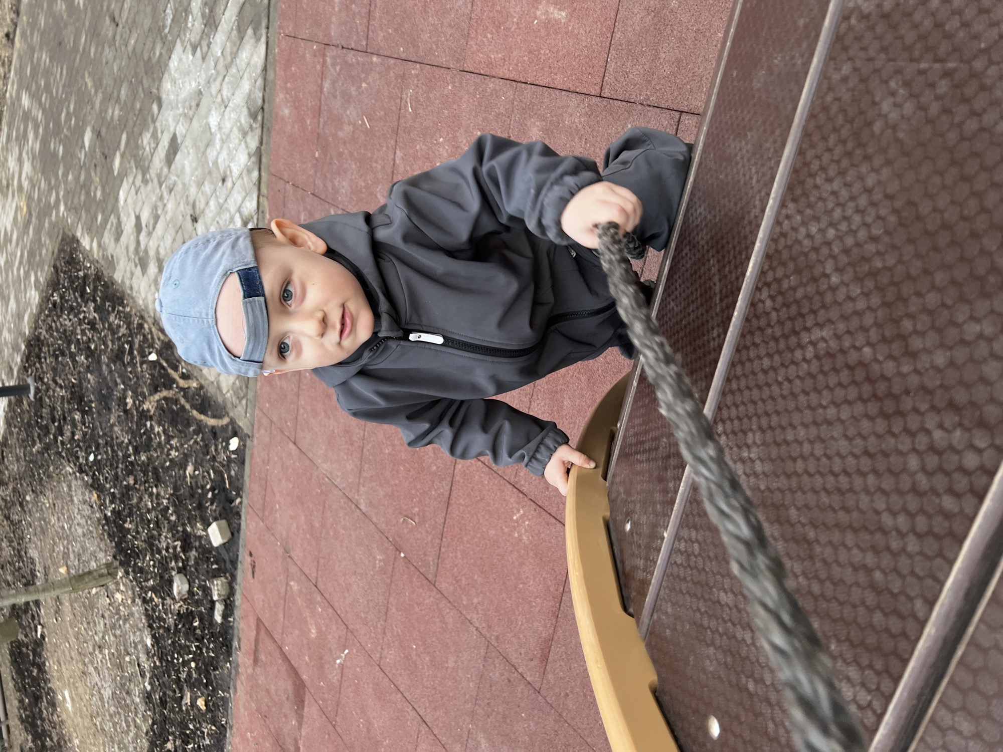 A young child wearing a grey jacket and a backwards blue baseball cap is climbing on a playground structure. He is gripping a grey rope with his right hand and holding onto a curved yellow support with his left. The structure appears to be made of brown material with a honeycomb pattern. The ground beneath the structure is covered in red tiles. To the left of the structure, there is a patch of land with dark soil and debris. The image suggests the location is likely a playground in Mytishchi, Russia.