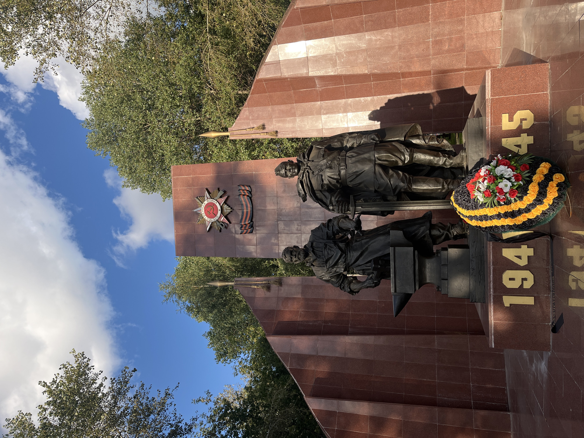 The image shows a monument with two bronze statues of men in military attire, standing in front of a red marble wall. The monument is inscribed with the numbers "1945" on a red marble base. A wreath with red, white, and yellow flowers and black and yellow ribbon is placed in front of the monument. The statues are in a park-like setting, with trees and a blue sky with white clouds in the background. This monument is the memorial dedicated to the Heroes of the Soviet Union, located in Moscow, Russia.