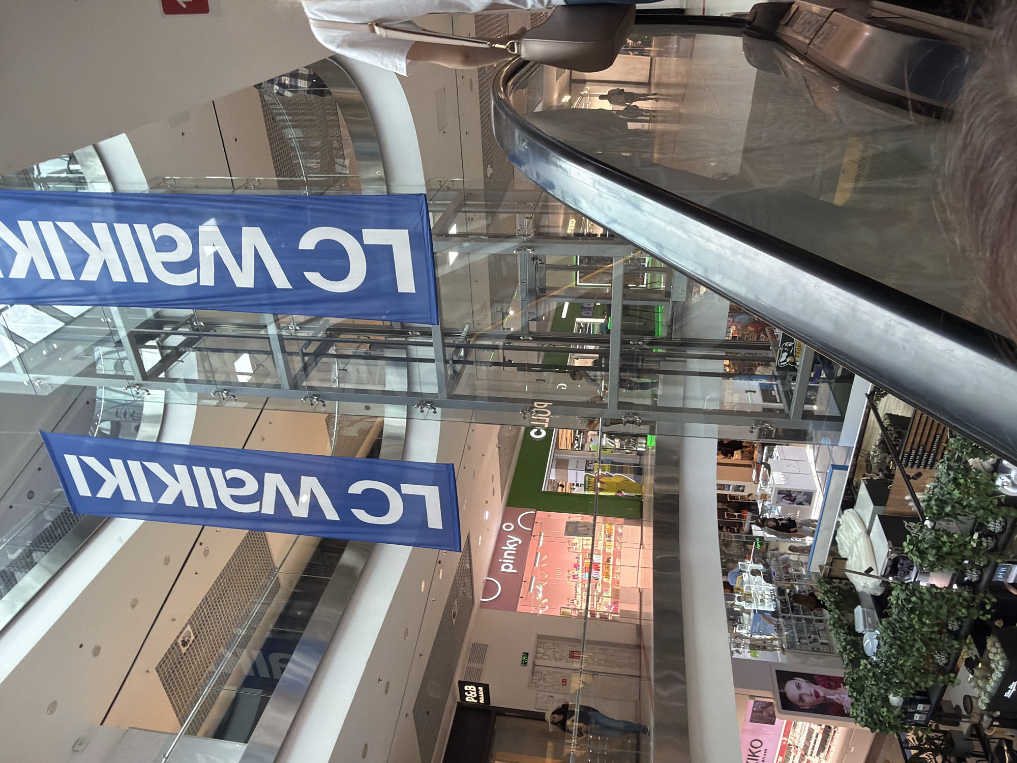 The image shows the interior of a modern shopping mall, likely in Cherkasy, Ukraine, characterized by multiple levels, glass railings, and escalators. Two large blue banners with white lettering, advertising "LC Waikiki," hang prominently in the central atrium. On the lower level, several storefronts are visible, including "pinky" and "P&B Pull&Bear." People are seen walking on the ground floor and on the balconies. An escalator ascends from the foreground, with the shoulder and head of a person visible at the top. The overall environment is brightly lit and suggests a busy commercial space.