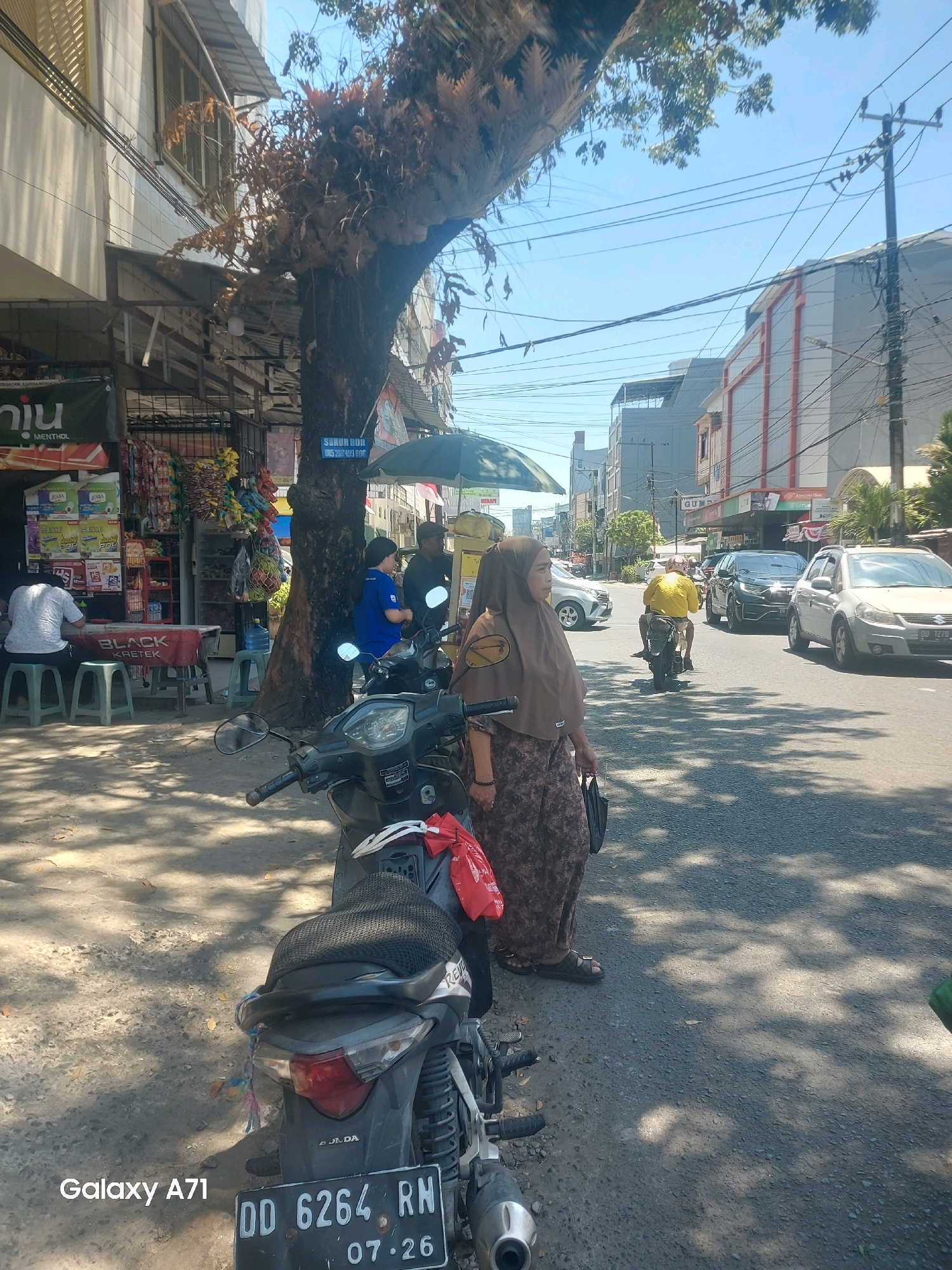 A street scene in Makassar, Indonesia features a woman wearing a brown hijab and patterned dress standing on a sidewalk next to a motorcycle. The motorcycle is partially visible in the foreground, with a red plastic bag hanging from its seat. Behind the woman, a tree with dry foliage grows prominently, casting shadows on the ground.
In the background, several buildings line the street, some appearing to be shops with awnings. A power pole with multiple wires is visible, and there are other vehicles on the road, including cars and motorcycles. One motorcycle with a rider in a yellow shirt is in motion. The sky is clear and blue. A street vendor stall with various goods is on the left side of the image. A blue sign with white text is attached to the tree. The license plate of the motorcycle in the foreground reads "DD 6264 RM 07.26". The bottom left corner of the image displays "Galaxy A71".