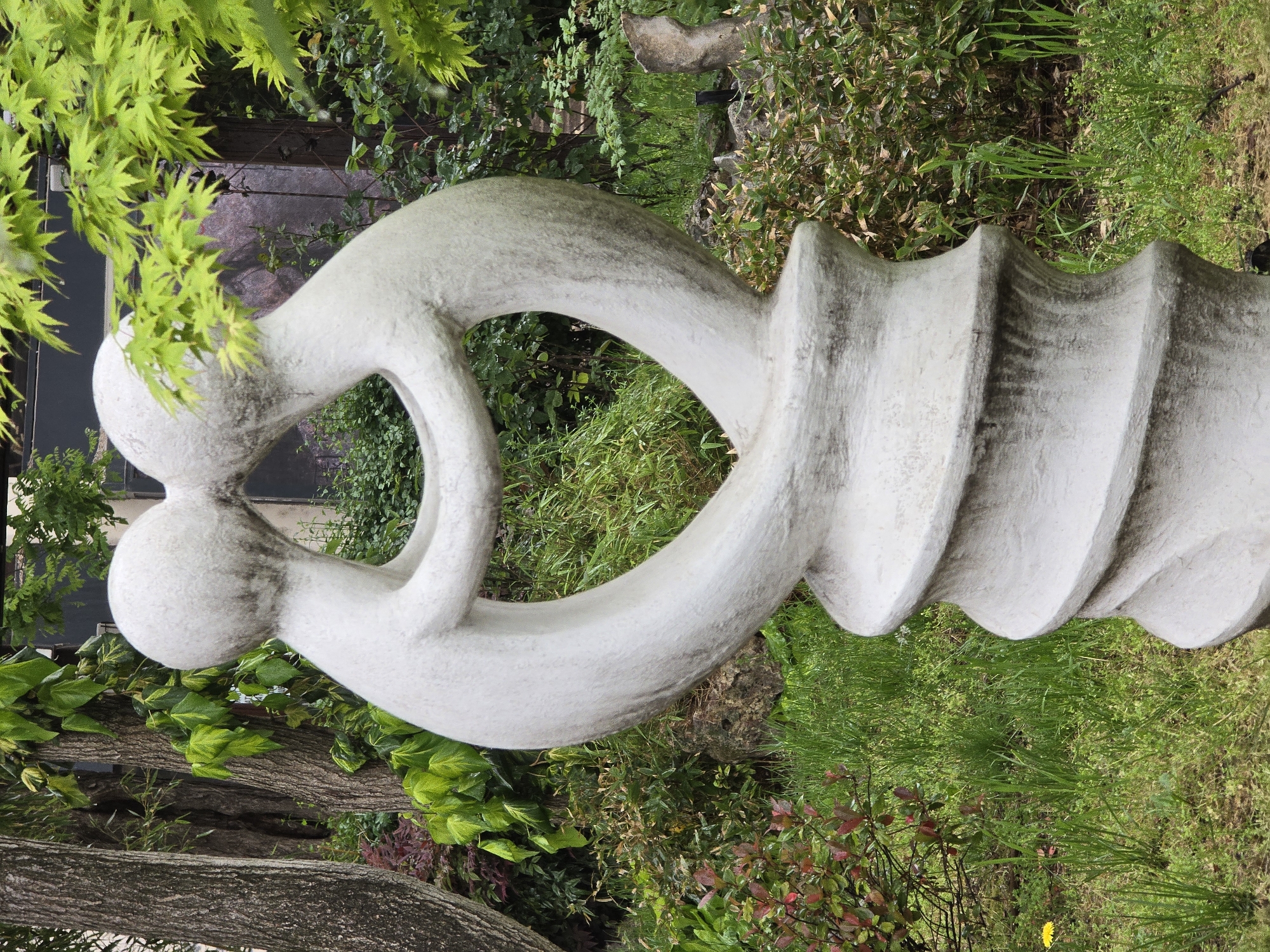 A gray stone sculpture stands in a grassy area in an unknown location. The sculpture features two intertwined, smooth spherical shapes transitioning into a screw-like form with ribbed undulations. The surrounding environment consists of grass, various types of greenery, and a tree trunk.