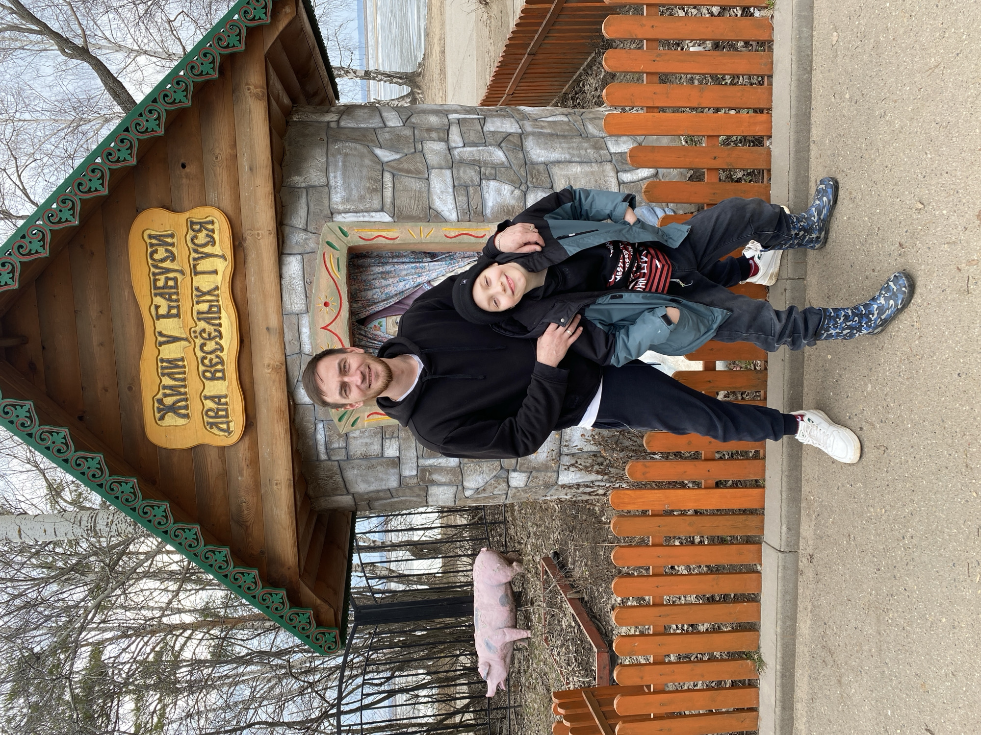 A man and a child, likely a boy, are captured in a heartwarming portrait outdoors in Chaikovsky, Russia. The man, wearing a black hoodie and dark pants, stands with his arm around the child, who is dressed in a black beanie, a blue-gray jacket, dark pants, and distinctive blue camouflage boots. Both are smiling directly at the camera, conveying happiness and affection.
They are positioned in front of a quaint, decorative wooden house with a stone-clad base. The house features a green, intricately carved trim along its roof eaves, and a small window with a colorful painted frame and a curtain visible inside. A prominent wooden sign hangs on the house, bearing text in Cyrillic: "ЖИЛИ У БАБУСИ ДВА ВЕСЕЛЫХ ГУСЯ," which translates to "GRANDMA HAD TWO JOLLY GEESE," referencing a popular Russian children's folk song.
To the right of the subjects, an orange wooden fence runs alongside a concrete pathway. Near the fence and behind the man, a pink pig statue is visible on the ground. The background reveals bare trees, indicative of late winter or early spring. Beyond the trees, a wide body of water, possibly a lake or river, appears to be covered with ice or slush, reinforcing the seasonal context. The sky is overcast, providing soft, diffused lighting. The overall scene suggests a pleasant daytime outing in a scenic, perhaps themed, park or recreational area.