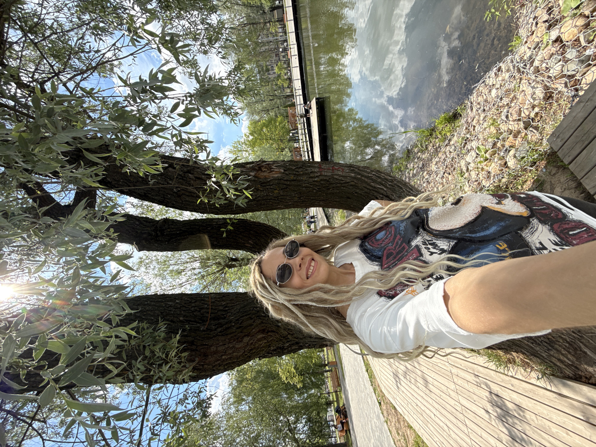 A vibrant, sunny outdoor selfie captures a woman with long blonde dreadlocks and round sunglasses smiling broadly at the camera. She is wearing a white t-shirt adorned with a colorful, abstract graphic design that includes various shapes and possibly some indistinct text. The image is taken from a low, tilted angle, with a bright sunburst and lens flare visible through the tree canopy in the upper left.
The setting is a park or natural area in Rusanovo, Russia, characterized by lush greenery and a body of water. Large tree trunks with rough bark and abundant green-silver foliage frame the left side of the image and extend overhead. To the right of the woman, a calm pond or lake reflects the clear blue sky with scattered white clouds, as well as the surrounding trees and distant structures. The water's edge features a gabion-like stone reinforcement structure held by wire mesh, beside a well-maintained wooden walkway or deck.
In the background, a paved path with park benches is visible, and the faint figures of other people can be seen. A wooden pier extends into the water in the distance. The strong sunlight indicates it is daytime, likely late morning or early afternoon, creating a bright and cheerful atmosphere. The woman appears happy and relaxed. No clearly legible text is visible other than the abstract design on her shirt.
