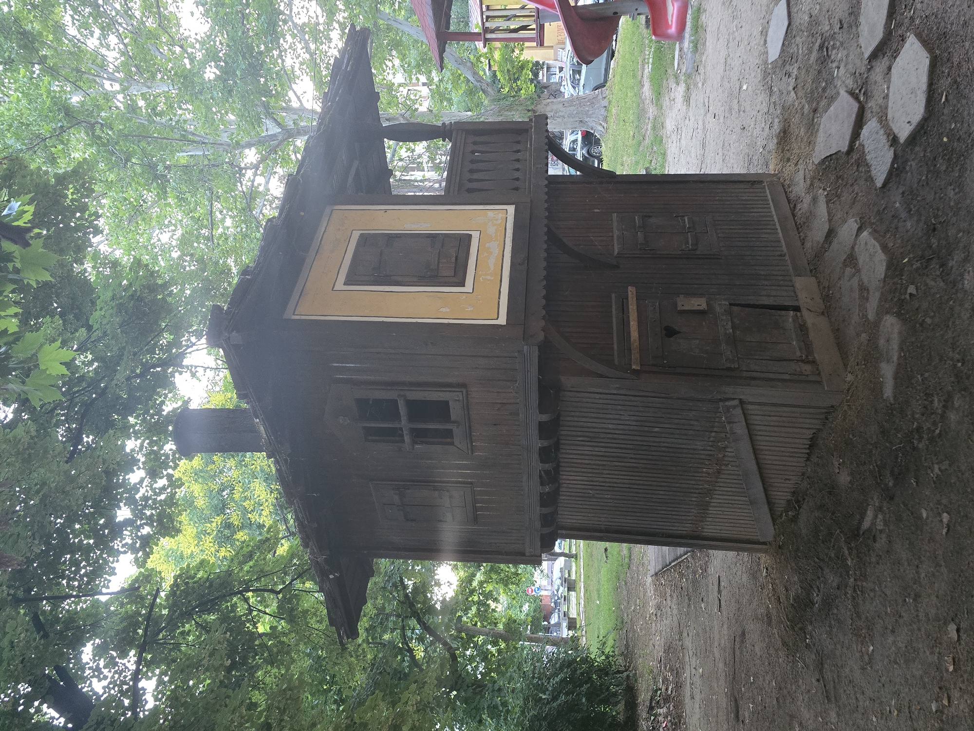 The image shows a weathered, two-story wooden playhouse located outdoors in a park-like setting. The playhouse is dark brown, with a slanted roof and a chimney. The upper story features a window with shutters and a small balcony with decorative railings. The lower story has two small doors and a window-like opening with a heart shape carved into it. The playhouse appears to be situated on a patch of dirt with scattered flat stones forming a rudimentary path. In the background, trees with lush green foliage dominate the scene, with sunlight filtering through the leaves. Beyond the trees, a street with cars and a red playground slide are partially visible, suggesting an urban or suburban environment. The scene is likely in Plovdiv, Bulgaria, given the context of the request.