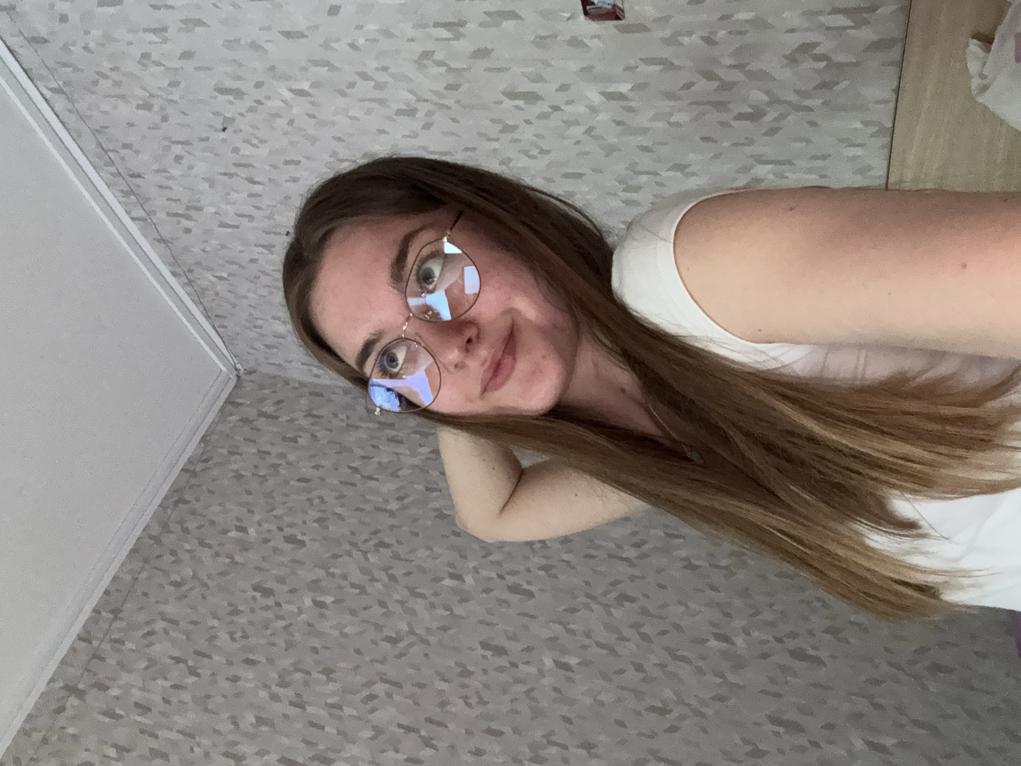 The image features a young woman, likely in her late teens or early twenties, captured in a significantly tilted frame. She has long, straight, light-to-medium brown hair that extends over her shoulder. She wears round, thin-framed glasses, and her eyes, visible through the lenses, show reflections of light sources. Her expression is calm, with a subtle smile. She is wearing a simple white short-sleeved top. Her right arm is bent at the elbow, with her hand possibly supporting her head or holding the camera.
The background consists primarily of a light grey patterned surface that resembles linoleum or carpet tiles, suggesting an indoor setting. In the upper left portion of the tilted image, a plain white wall is visible, meeting the patterned surface at an angle, indicating an interior corner. There are no clear indications of time of day or weather, as the lighting appears to be internal. No discernible text is visible anywhere in the image. The overall impression is an informal portrait, possibly a selfie, taken from a low angle while the subject is reclining or lying down, given the perspective. This scene takes place in Perm, Russia.