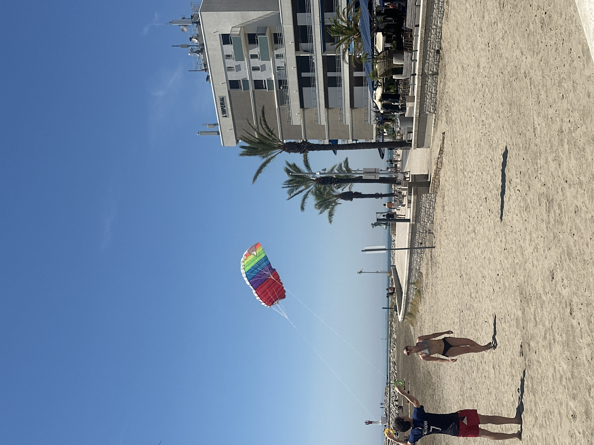 The image captures a sunny day at a beach, likely in the late morning or early afternoon given the bright sunlight and long shadows. The scene is set against a backdrop of a modern building, possibly a hotel or resort, with "L'IMPERIAL" visible in large letters on its side. Palm trees dot the landscape, suggesting a warm climate.
In the foreground, two individuals are engaged in flying a colorful, rainbow-patterned kite. One person is holding the kite strings, seemingly controlling its movement in the clear blue sky. Another person, wearing a bikini, is walking on the sandy beach, casting a distinct shadow. Further down the beach, another person in red shorts and a blue t-shirt is also visible. The presence of beach chairs and umbrellas suggests a leisurely atmosphere. The sea is visible in the distance, adding to the coastal setting. The overall mood appears relaxed and enjoyable, indicative of a pleasant day outdoors.