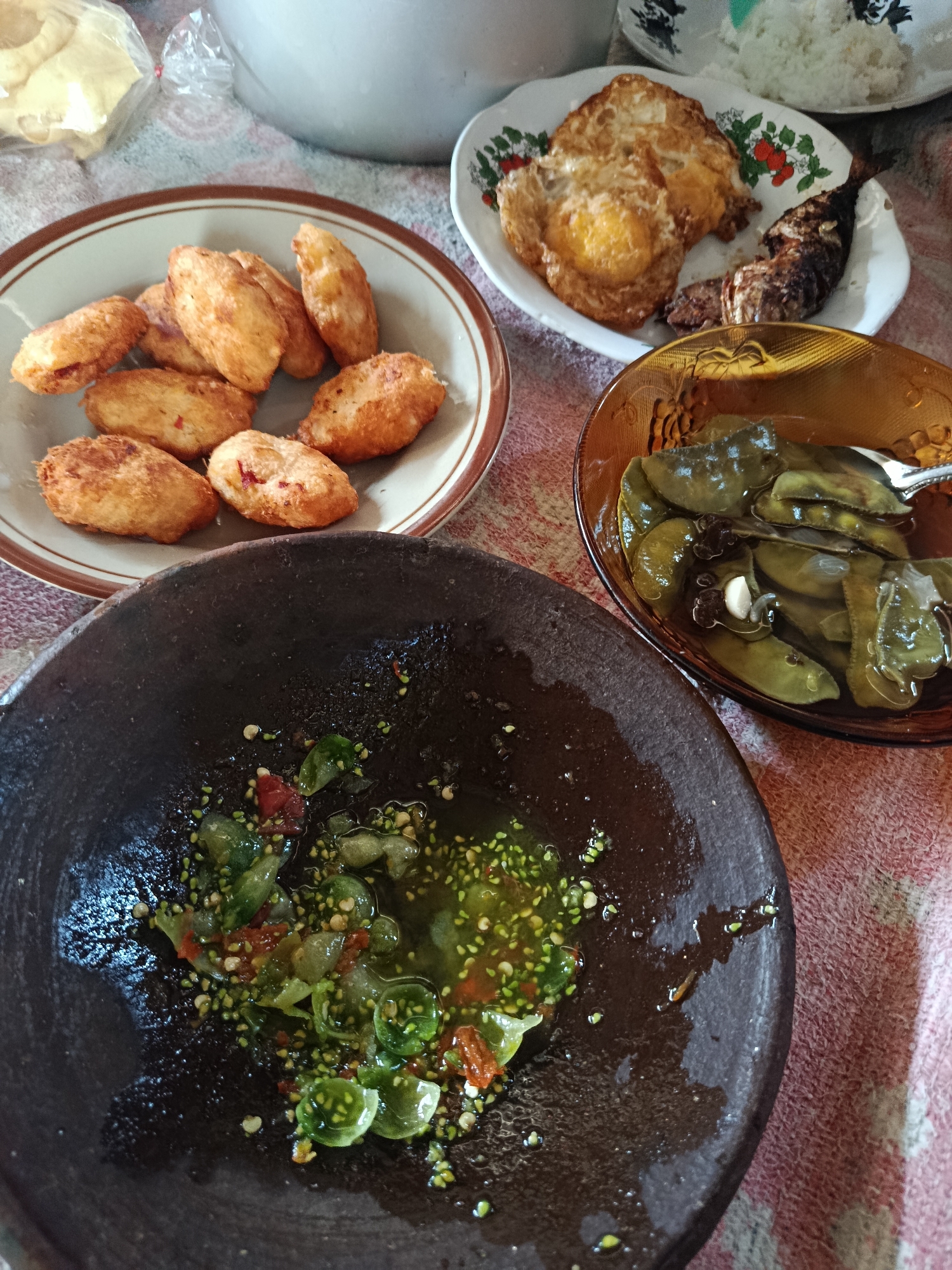 An overhead, eye-level shot shows a meal laid out on a red and white checkered tablecloth. In the foreground, a dark, stone mortar holds a spicy green chili sauce with visible chunks of chili and seeds. To the upper left, a light brown plate contains several golden-brown fried potato fritters. To the upper right, a white plate features a fried egg and a grilled fish. A clear amber bowl to the right holds pieces of cooked green beans in a light sauce. In the background, a white rice bowl and a bag of snacks are visible. The setting appears to be a home dining table in Lamongan, Indonesia, possibly for lunch.