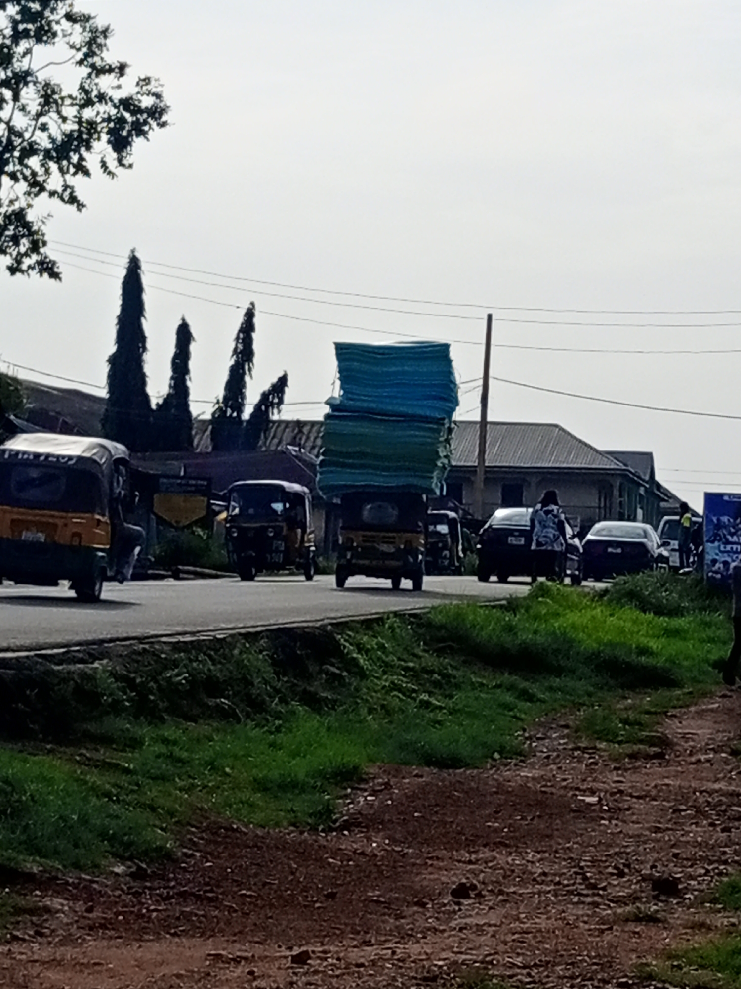 The image shows a street scene on Rukuba Road, Nigeria. In the foreground, there is a dirt and grass embankment. The road itself is visible, with several vehicles on it. On the left, a yellow auto-rickshaw is parked, with a person visible inside. Further down the road, another auto-rickshaw, also yellow, is moving, carrying a large, stacked load of blue and green material. Other vehicles, including cars and more auto-rickshaws, are present on the road, some of which are also carrying loads. In the background, there are buildings with pitched roofs and tall, slender trees. The sky is overcast.