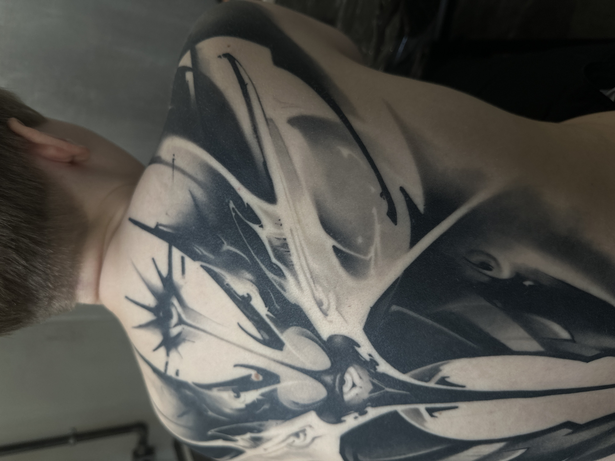 The image depicts the back and part of the upper arm of a person with a complex, abstract black and gray tattoo. The tattoo appears to cover a significant portion of the back and extends onto the arm. The background is out of focus and appears to be a wall and possibly a piece of equipment. The focus is on the tattoo and the skin it is on. The location is not discernible from the image.