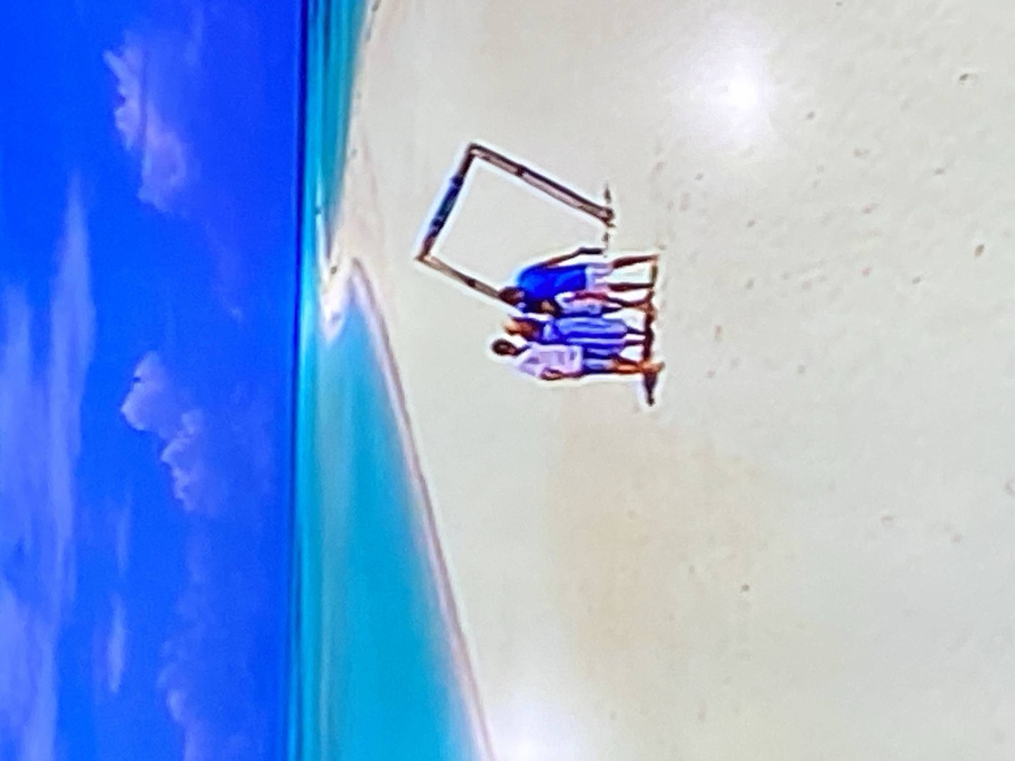 The image depicts a serene beach scene with turquoise water and a bright blue sky. In the foreground, a wooden frame stands on the sand, tilted at an angle. Three people are gathered near the frame. A man in a blue shirt and white shorts stands to the right of the frame, facing it. To the left of the frame, a couple stands together, with the man wearing a white shirt and striped shorts, and the woman in a blue and white striped dress. A small child, possibly a girl, is standing between them, also wearing a blue and white striped dress. The sand is light-colored, and the water appears calm. The image is somewhat pixelated, making it difficult to discern fine details. The location is not Yekaterinburg, Russia; it is a tropical beach.