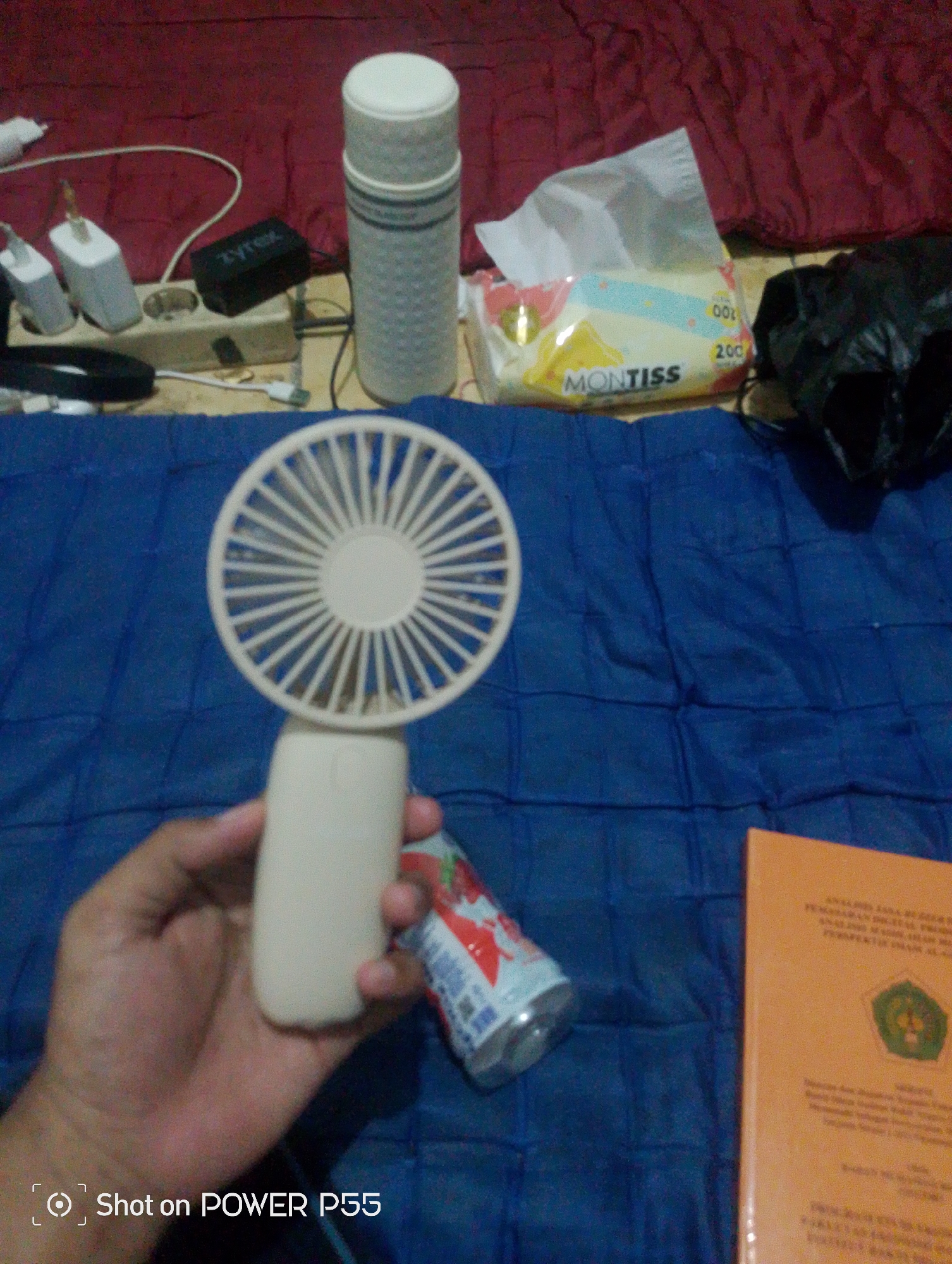 The image depicts a close-up, low-angle view of a person's hand holding a small, beige, portable fan. The fan has a circular, white, plastic grill. To the left of the fan, partially obscured by the hand, is a partially consumed beverage bottle with a red and white label. In the background, on a blue surface, is an orange book with text and a green emblem. Further back, on a red surface, are several objects including a power strip with electrical adapters, a black adapter labeled "Zyrex", a roll of tissues with the brand "MONTISS", and a black plastic bag. The scene appears to be indoors, possibly a bedroom, given the red fabric background which resembles a blanket or bedspread. The image includes a watermark in the bottom left corner that reads "Shot on POWER P55". The overall impression is that of a casual, personal setting.