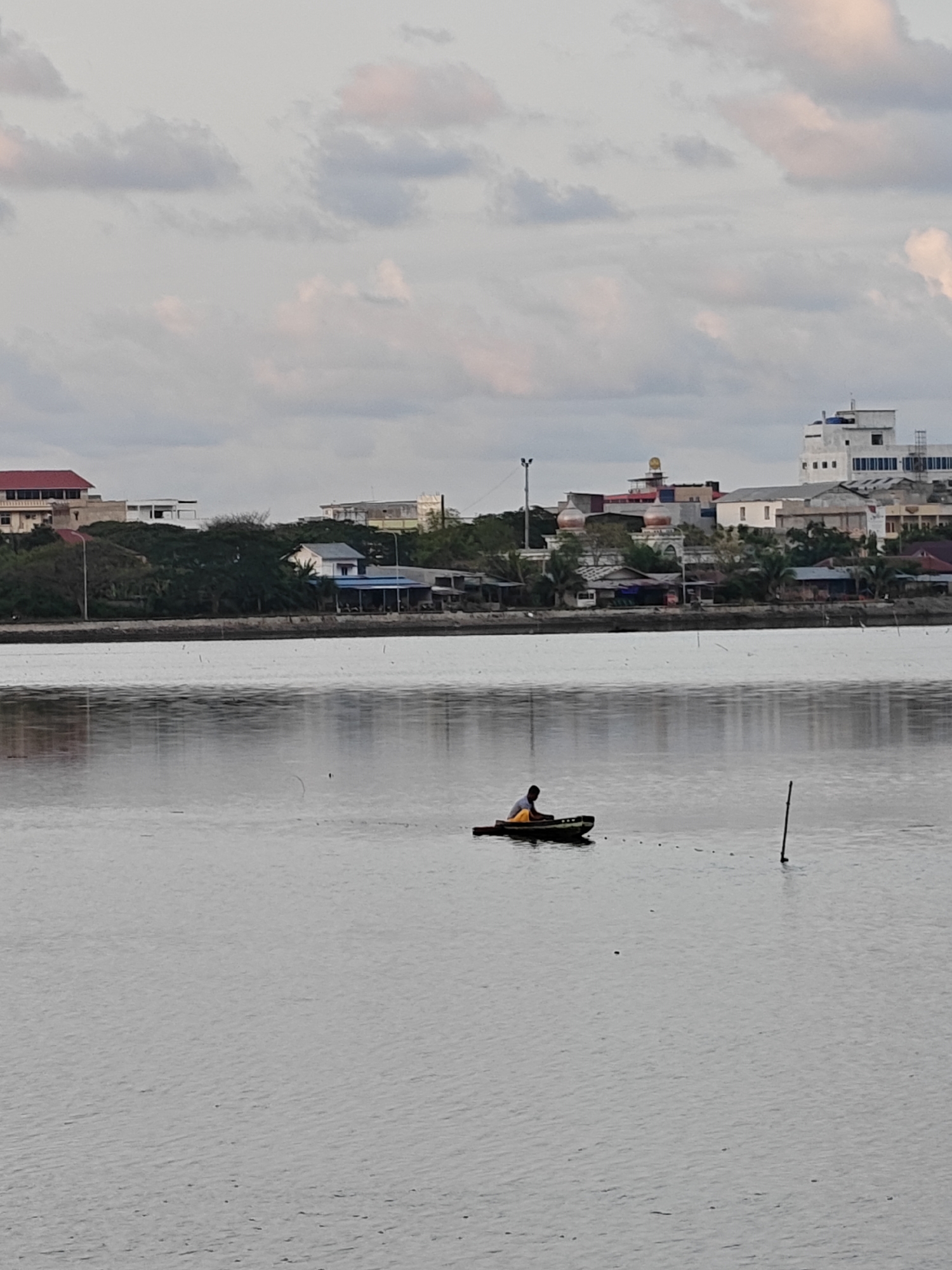 An eye-level outdoor photograph shows a man fishing in a small boat on a grey body of water. The man is centered in the image horizontally. He appears to be using a fishing net, attached to a line extending from his boat to a pole standing in the water. Buildings are in the background, including a white high-rise, a structure with two white domes (likely a mosque), and other residential buildings and commercial establishments. Trees are visible on the shoreline. The sky is filled with grey and white clouds. The location is near the city of Lhokseumawe, Indonesia.