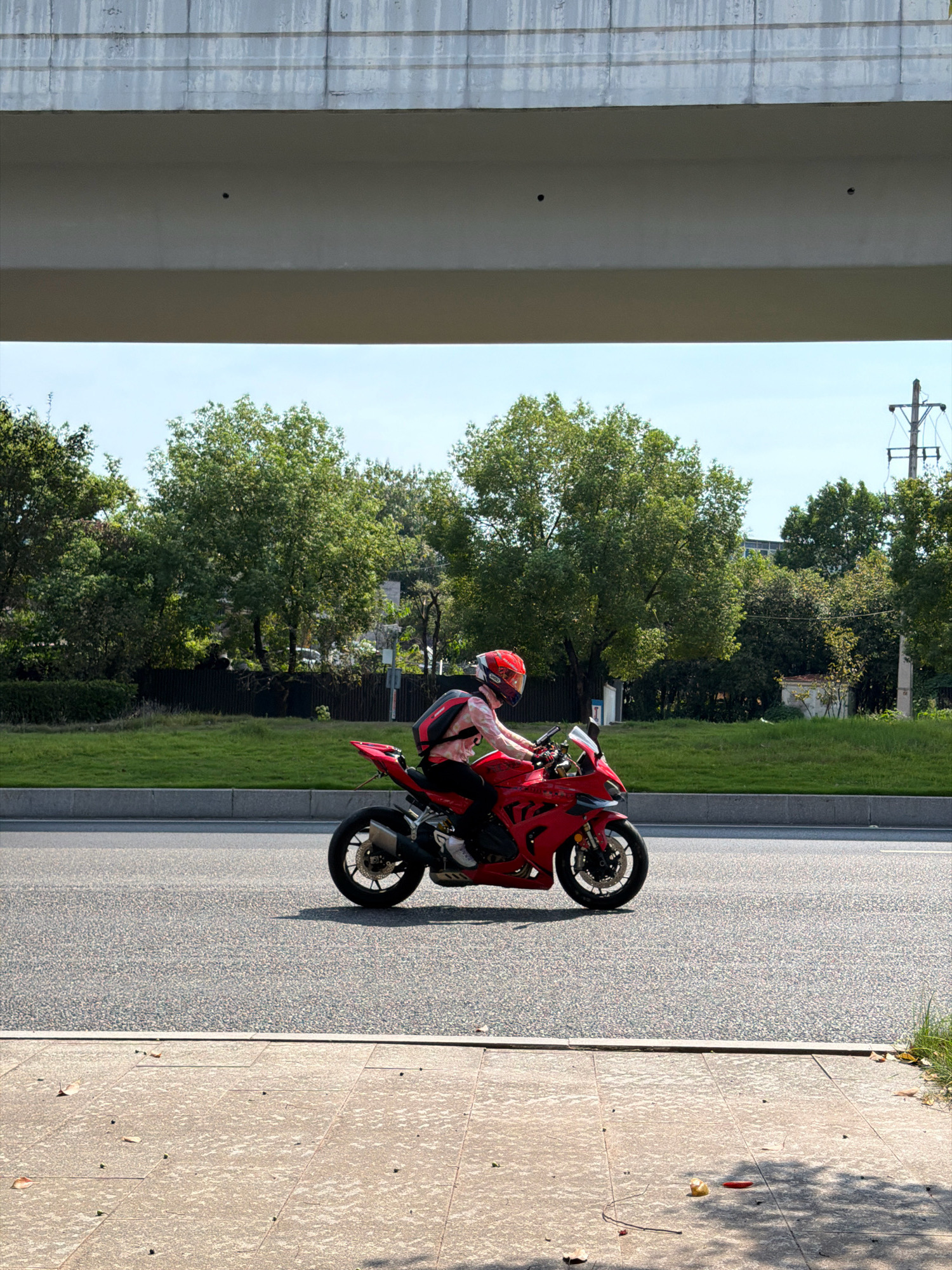 A person wearing a red helmet rides a red sport motorcycle along a paved road, with a sidewalk in the foreground and a grassy embankment and trees in the background. The motorcycle has a silver exhaust pipe and is angled slightly towards the viewer. The person's clothing is partially visible, with a red and black backpack worn over a light-colored shirt. The scene appears to be set on a bright, sunny day, with a portion of an overpass visible in the upper part of the frame. The location is confirmed to be Jiangjin District, Chongqing, China.