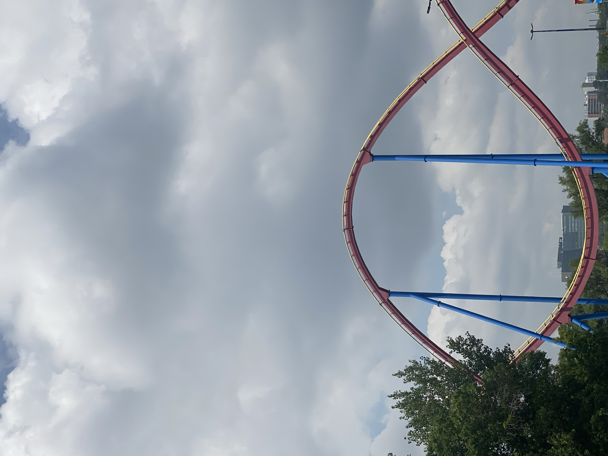The image captures a roller coaster track with red rails and yellow accents, supported by blue pillars, set against a cloudy sky. The track features a distinct loop, suggesting it's part of an amusement park. A lush green tree is visible on the left side of the frame. In the background, several buildings, including what appear to be office buildings or apartment complexes, are present, indicating an urban or suburban setting. The roller coaster is located at La Ronde amusement park in Longueuil, Canada. There are no people visible in the image, and no specific actions or interactions are depicted.