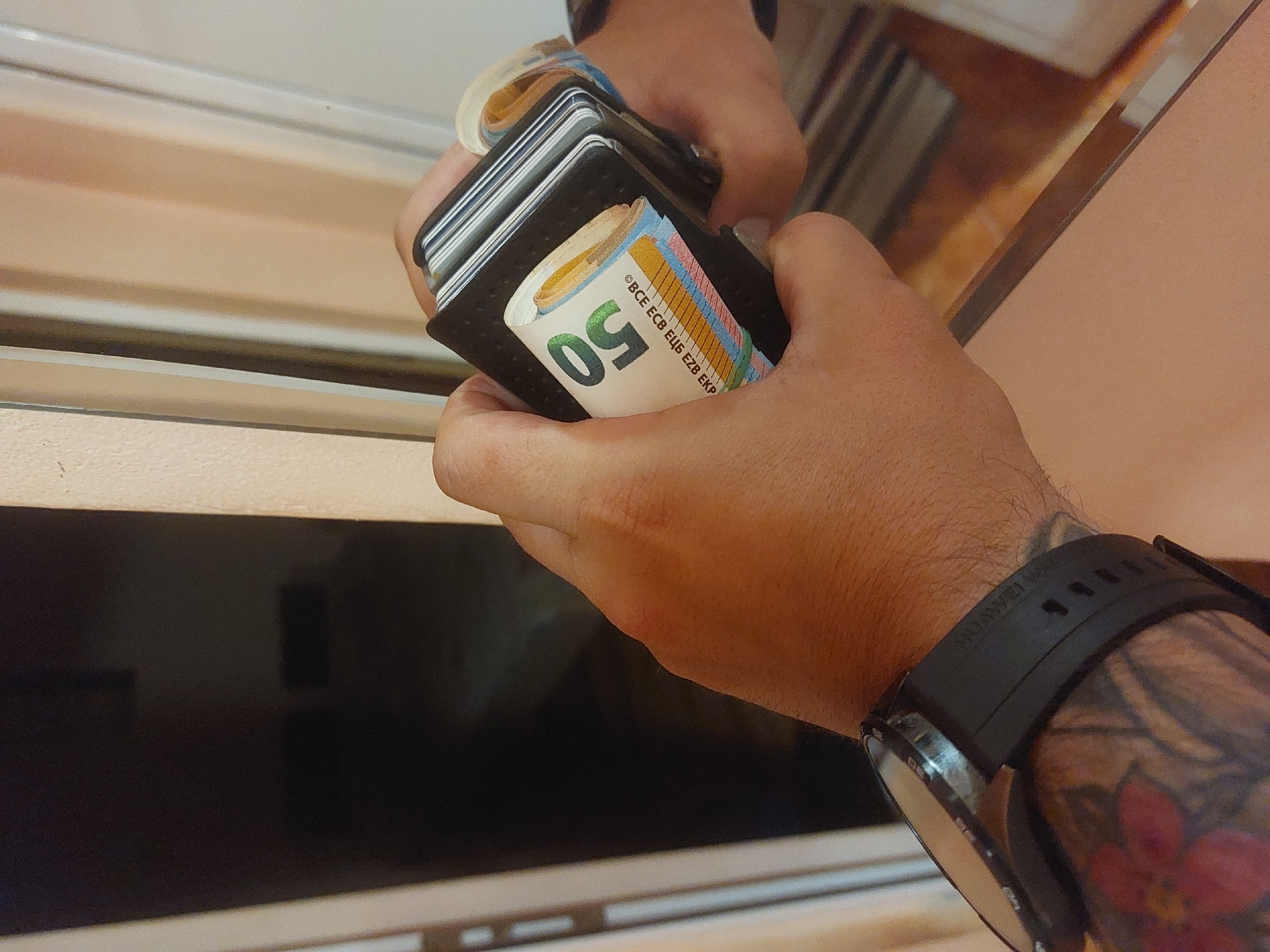 A person is holding a wallet filled with euro banknotes in their hands. The wallet is black and holds several cards as well as the rolled-up bills. The foreground hand has a visible watch and a tattoo on the wrist. The background is blurred, but it appears to be an interior space, possibly near a window or reflective surface. No information about the location in the city of Tsarevo, Bulgaria is available in the image.
