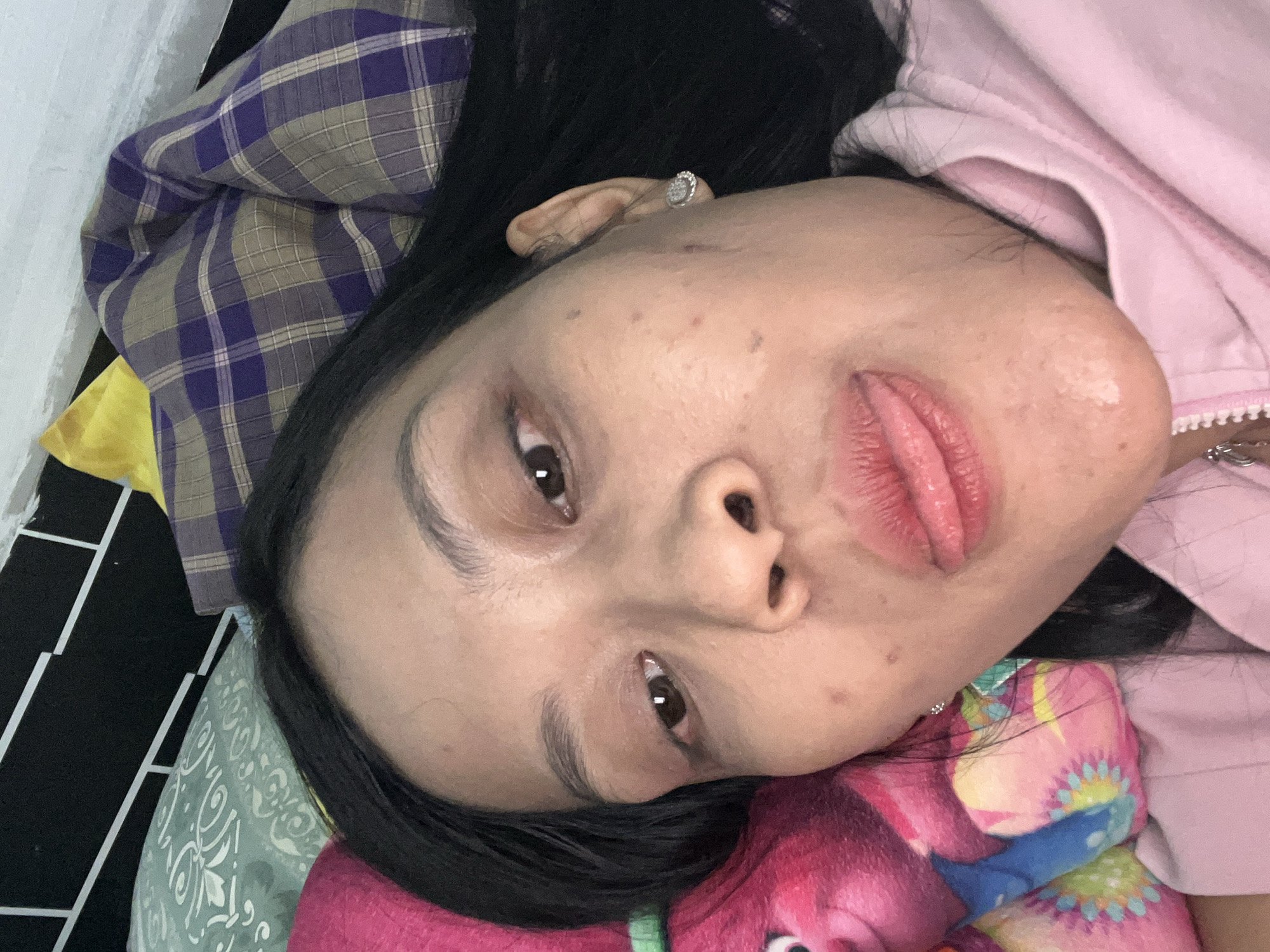 A close-up, selfie-style photograph features a young woman with long, dark hair lying down. Her face is the primary subject, with her lips slightly parted and her tongue visible. She is wearing a pink hooded jacket. To her left, a pink stuffed toy, possibly a character from the movie Trolls, is visible. The background consists of pillows and bedding with various patterns, including plaid and floral designs. The lighting appears to be artificial, casting a soft glow on her face. The image is taken indoors, likely in a bedroom, and there are no discernible landmarks or details that would indicate a specific location within Yogyakarta, Indonesia. The woman's skin has a few minor blemishes and her eyebrows are lightly filled in. She is wearing a small, sparkly earring in her right ear. The overall impression is a casual, personal moment captured in a relaxed domestic setting.