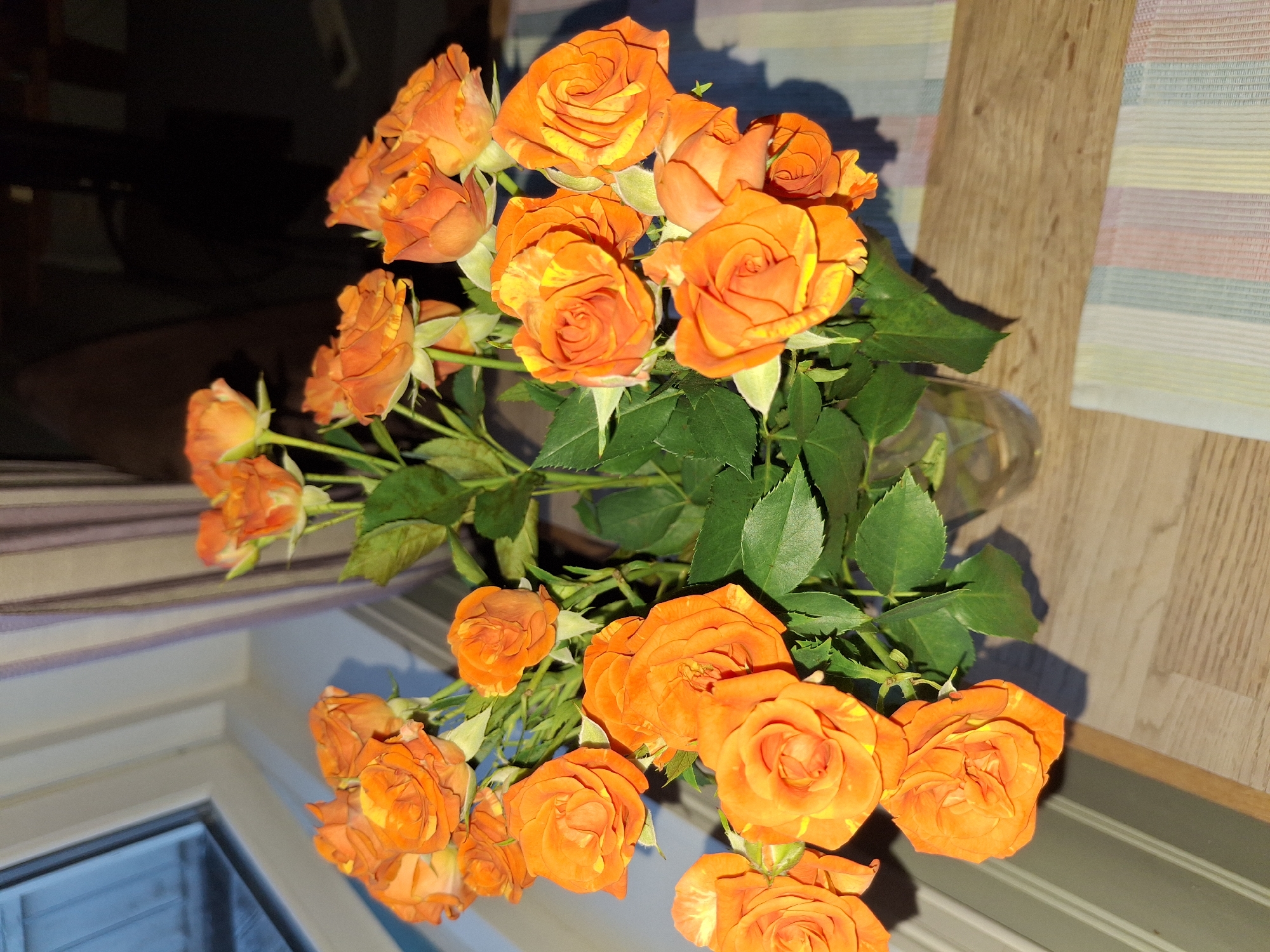 A bouquet of bright orange roses with yellow streaks is displayed in a clear glass vase. The vase sits on a wooden surface, possibly a table, covered in part by a striped placemat. In the background, a window with white trim is visible, revealing a glimpse of the outdoors. Behind the flowers, a dark, out-of-focus background suggests an indoor setting. The lighting indicates it is likely daytime. The scene is static and does not depict any actions or interactions. The location is identified as Åsgårdstrand, Norway.