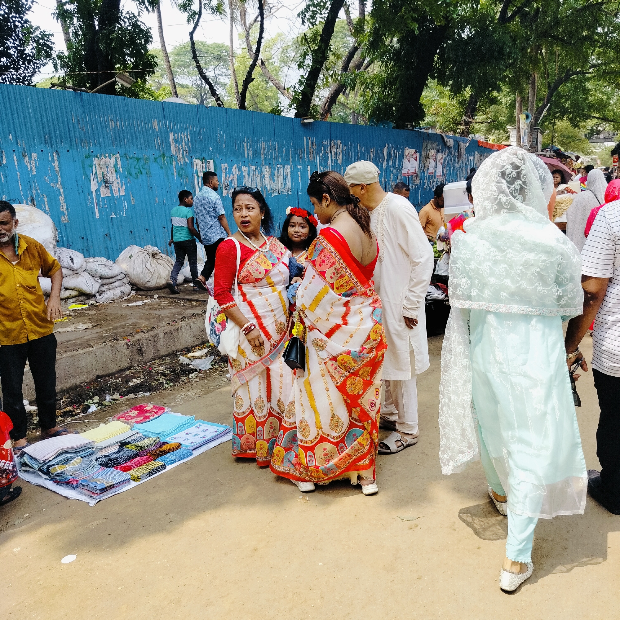 Captured in the urban landscape of Dhaka, Bangladesh, the image portrays a bustling street scene. A group of individuals are positioned near a sidewalk adjacent to a weathered blue fence, behind which trees are visible. Two women stand out with their traditional saris, one with red and white patterns, and the other with similar designs. A man in white traditional attire stands near them. A young girl with a flower crown adorns their ensemble. To the left, a man in a yellow shirt stands beside a makeshift display of colorful textiles laid out on the ground. The sidewalk is littered with debris, and white sacks are piled against the fence. Passersby are seen in the background, adding to the vibrant street life.