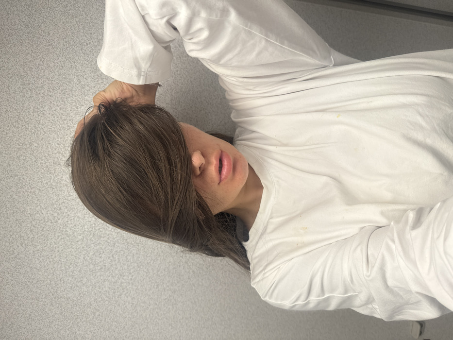 A person with dark brown hair covering their forehead lies on a textured, light gray surface. They wear a long-sleeved white shirt. Their hand rests on their head. The face is partially obscured by hair, revealing the mouth, nose, and part of the cheek. The lighting appears soft and diffused. Several stains are present on the shirt. The image is rotated counter-clockwise.