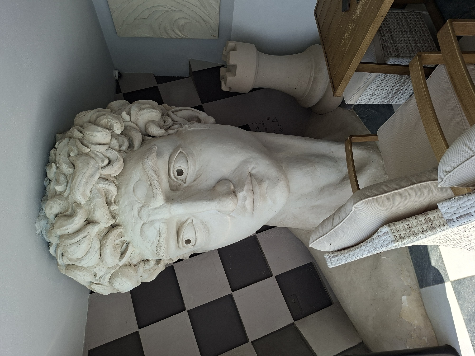 The image depicts an indoor scene with a colossal statue of a male head, resembling Michelangelo's David, dominating the frame. The statue is a pale, off-white color and has a textured, unfinished appearance, particularly in the curly hair. The head is so large that it appears to emerge from the wall and ceiling. To the left of the statue, a black and white checkered pattern covers the wall. To the right of the statue's head, a large white chess rook sculpture stands. In the foreground, two light-colored, upholstered chairs with wooden frames are positioned in front of a wooden table. The overall setting suggests an interior design with a classical and game-themed aesthetic, potentially a cafe or a unique decorative space. The provided metadata indicates the location is Costești, Moldova. No people are visible in the image.