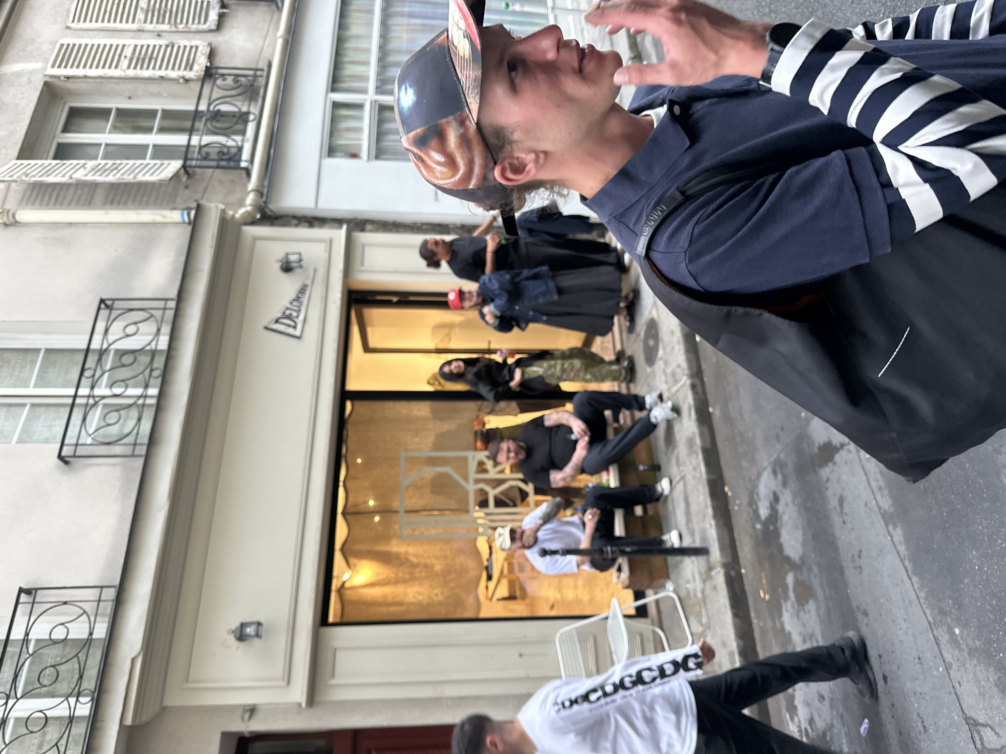 The image captures a street scene in what appears to be Paris, France, showing a mix of people standing, seated, and walking near a storefront. The overall composition is taken from an upward angle, making the ground appear on the right side of the frame.
The most prominent subject is a young man in the foreground, facing right (as per the rotated image) and looking slightly upwards. He wears a navy blue polo shirt over a white and black horizontally striped long-sleeve shirt, and a baseball cap featuring a striking graphic print, possibly of a face or abstract art. His left arm, adorned with a watch, is visible, and his right hand is raised near his chin, suggesting he is speaking or gesturing. A dark bag strap crosses his shoulder. On his polo shirt sleeve, a small black tag with "MMW" is visible.
In the mid-ground, a storefront with a large glass window is visible. Inside and immediately outside, several individuals are gathered. Two men are seated on white outdoor chairs in front of the shop. One wears a white long-sleeve shirt with repeating text, while the other is in a black t-shirt and dark pants. Further inside the store, other figures can be seen, including a person in a red cap, another with long dark hair, and one wearing camouflage-patterned pants.
The buildings lining the street exhibit classic Parisian architectural elements, with white walls, multi-paned windows, and decorative black wrought-iron railings. Some windows feature white louvered shutters. A white sign above the storefront entrance displays the partial word "DELORTUN". The pavement in the foreground appears damp in areas, suggesting recent rain.
In the bottom left corner of the image, the back of a man is visible, walking away. He wears a white long-sleeved shirt with the repeating text "CDGCDG" or "CDG DGC DCDG". The scene suggests a casual daytime gathering outside a boutique or café, with people engaged in conversation or waiting.