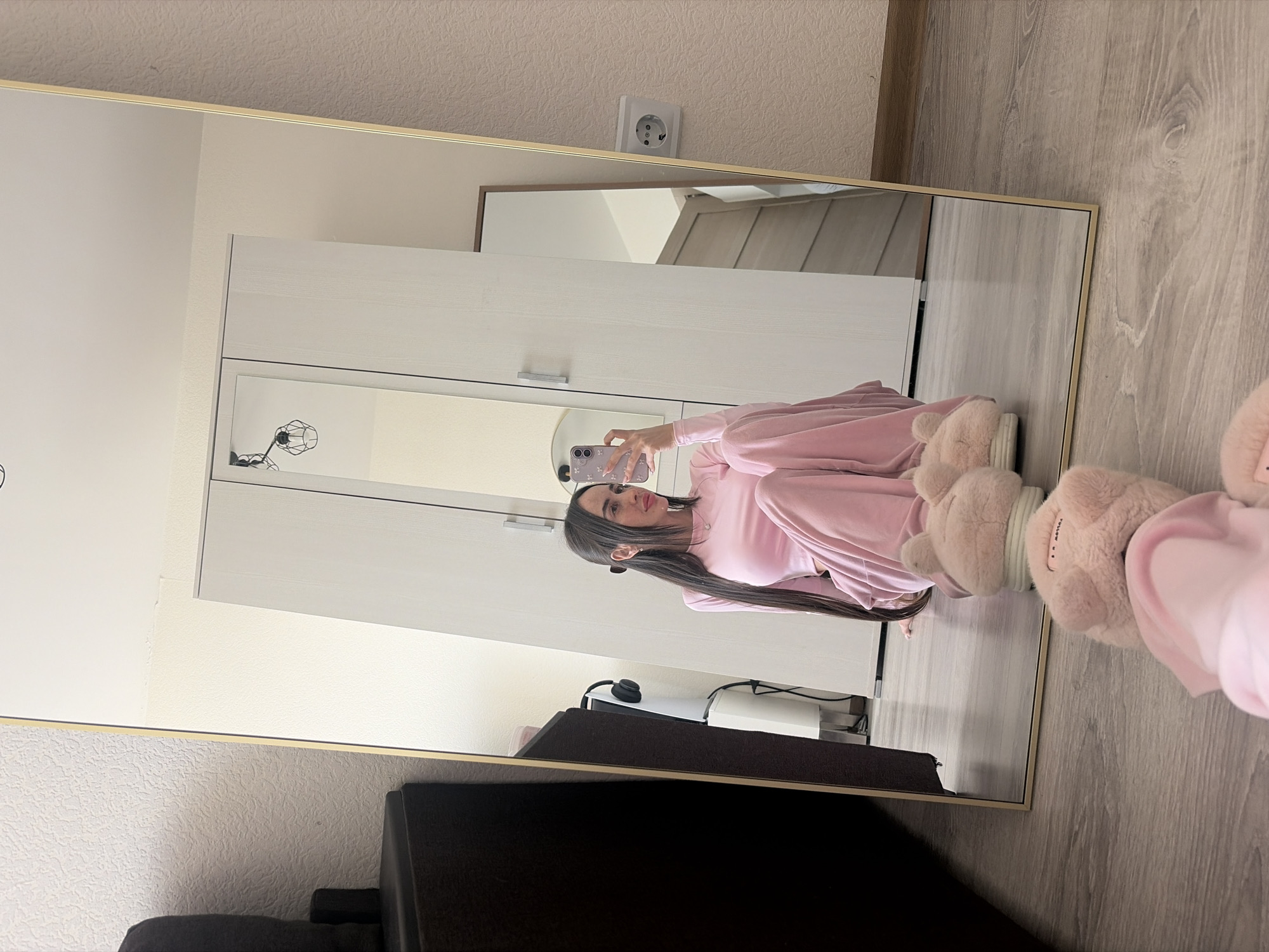 A woman is taking a selfie in a mirror inside a building. The location appears to be indoors with a wardrobe, a desk, and a power outlet visible in the background. The woman is wearing a pink outfit and fuzzy slippers. The photo is likely taken in Russia, based on the design of the power outlet, which is commonly used in Saint Petersburg, Russia.