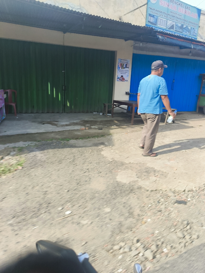The image shows a man walking on a dirt road in front of a shop. The man is wearing a blue shirt and tan pants. He is carrying a bottle of water in his right hand. The shop has green metal roll-up doors and a blue door with a sign above it that says "Bengkel Las Sinar Berkah". There is a table and chairs outside the shop. The image was taken in Jatiluhur, Indonesia. The coordinates are -6.57 latitude and 107.438 longitude. The image was taken from the perspective of a vehicle.