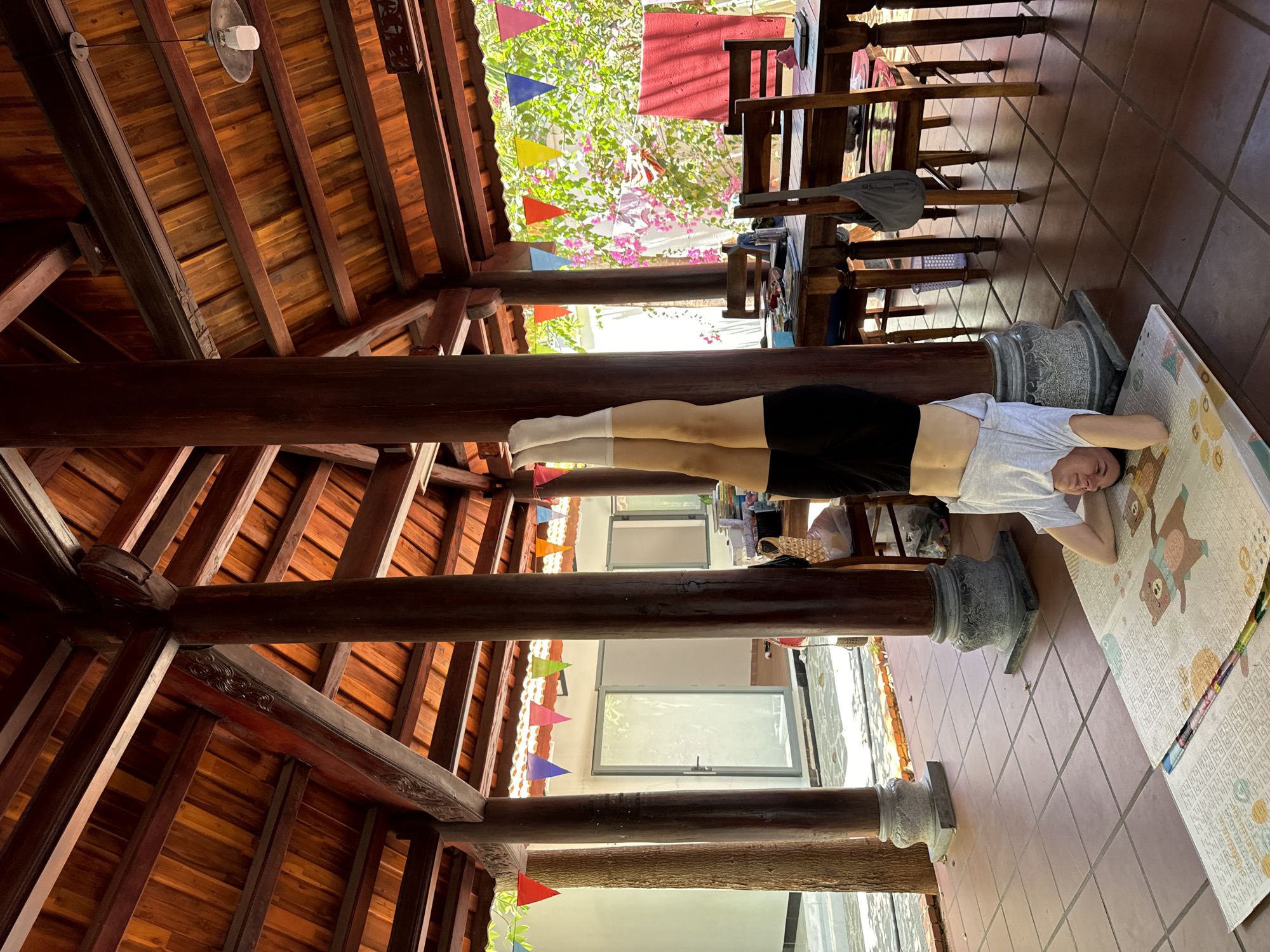 A woman is lying on a mat on a tiled floor. The scene is in a room with a wooden roof structure supported by wooden pillars with decorative concrete bases. Outside, visible through openings between the pillars, are hanging decorative banners and lush greenery. The location is Mũi Né, Vietnam.