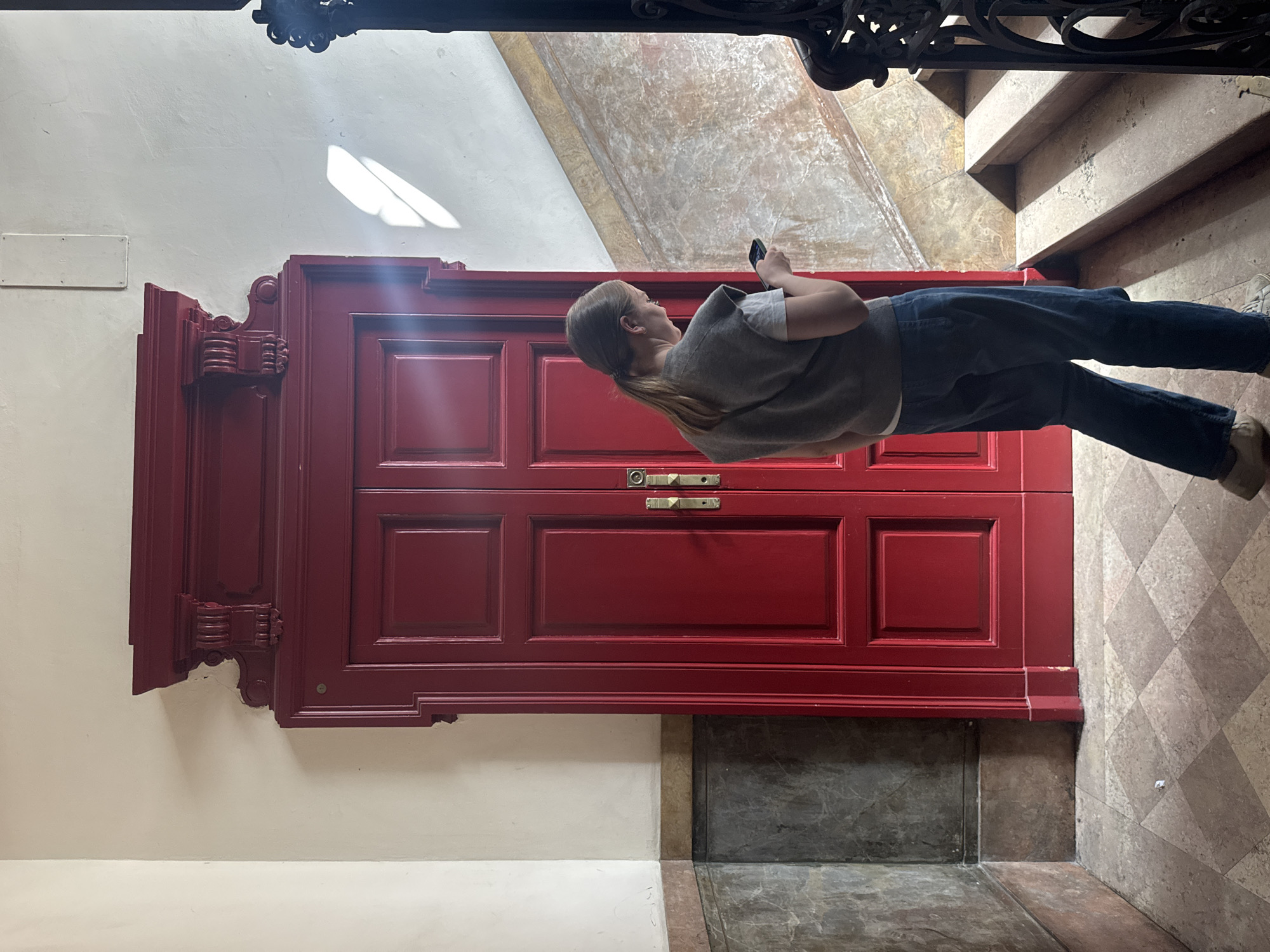 A medium shot captures a woman standing in an interior hallway or entrance area, facing a grand, ornate red double door. The woman has light hair tied in a ponytail and is dressed in a light grey short-sleeved top, a darker grey sleeveless vest, dark blue jeans, and light-colored shoes. She is holding a smartphone in her hands, looking slightly upwards as if observing or photographing something with her device.
The central subject is a vibrant, deep red double door, richly decorated with intricate moldings, raised panels, and prominent brass or gold-colored door handles and a lock. To the left of the door, a plain white wall features a small, blank rectangular plaque. A bright, rectangular patch of sunlight is cast on the upper section of this white wall, indicating daytime.
The floor in front of the door transitions from a dark, mottled grey and brown stone or tile pattern to a lighter, diamond-patterned floor surface extending towards a visible staircase on the right. The stairs are also made of a similar stone material. At the top edge of the image, a dark, decorative wrought-iron railing is partially visible, suggesting an upper floor or balcony. The overall setting implies a historical or well-maintained building, consistent with the architectural style often found in Milan, Italy. No legible text is visible on the plaque or elsewhere.