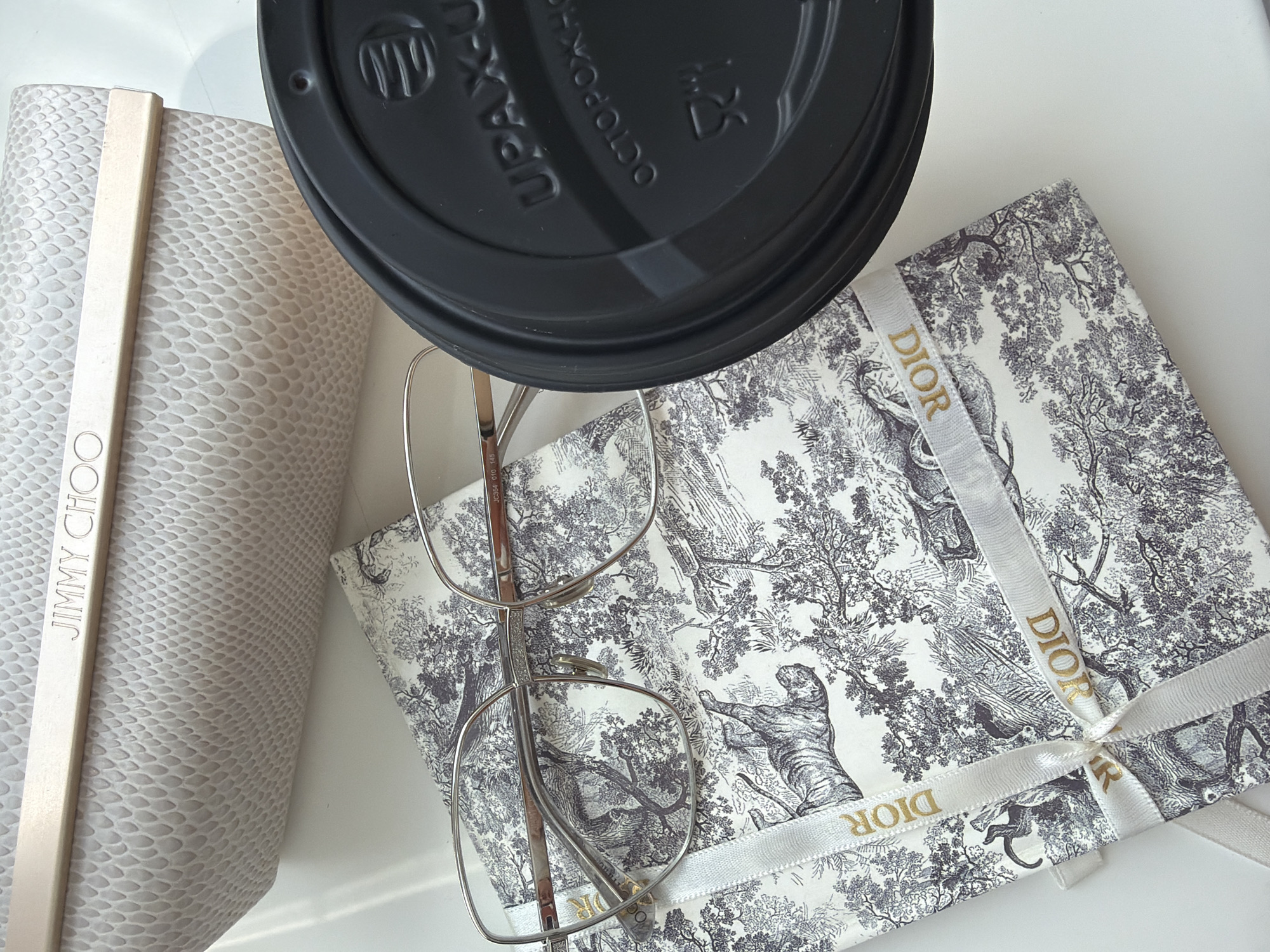 A high-angle shot captures a collection of items arranged on a white surface, likely indoors. At the left, a light-colored case embossed with a snakeskin texture and bearing the brand name "JIMMY CHOO" in a vertical gold strip is positioned. In the center, a black disposable coffee cup lid dominates, partially obscuring the objects beneath. A pair of silver eyeglasses with thin metal frames sits askew, partially covering the cup and the box below. To the right, a Dior-branded box wrapped in a white ribbon printed with the word "DIOR" in gold is visible. The box features a black-and-white toile-like pattern depicting various wild animals and landscapes. The overall arrangement suggests a casual and possibly transient setting, such as a desk or tabletop.