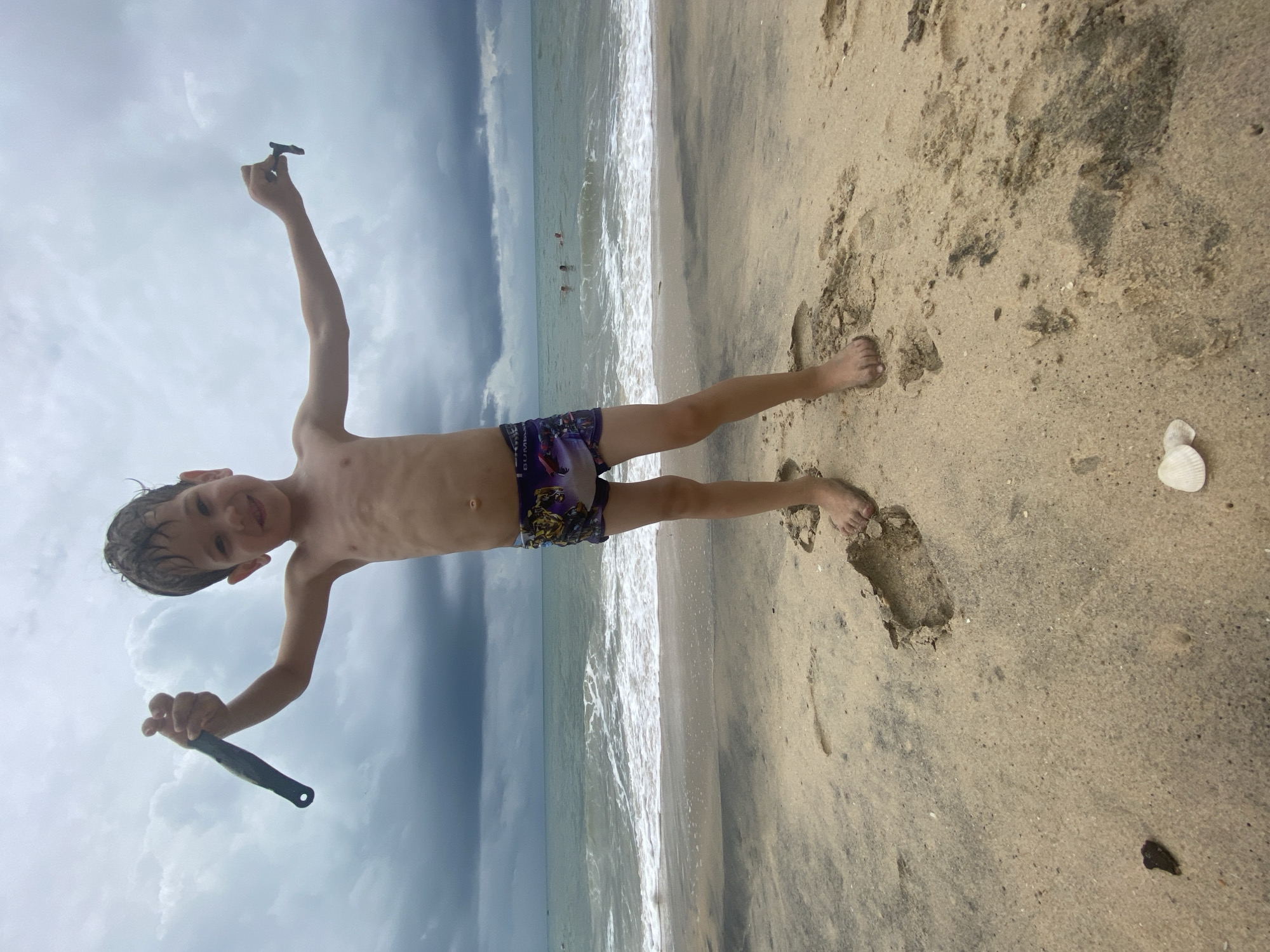The image depicts a young boy standing on a sandy beach, against a backdrop of the ocean and a cloudy sky. The boy, appearing to be between 5 and 8 years old, is fair-skinned with wet, dark hair and is shirtless, wearing colorful swim trunks featuring indistinct characters and text/logos, possibly "BUMBLEBEE" or a similar brand, on his left thigh. He stands with his arms outstretched, holding a small, dark object in each hand—one resembling a miniature shovel or tool, the other a stone or shell. He is smiling directly at the camera, conveying a sense of happiness and playfulness.
The setting is a beach in Phu Thuy Ward, Vietnam. In the midground, the gentle waves of the ocean break on the shore. Two other individuals are visible in the distance, partially submerged in the water. The sky above the ocean is overcast with dense clouds, suggesting a cloudy day or possibly an impending storm. The sand in the foreground shows numerous footprints, and a distinct white seashell lies near the bottom right. The overall atmosphere is that of a playful moment during a beach outing, despite the cloudy weather.