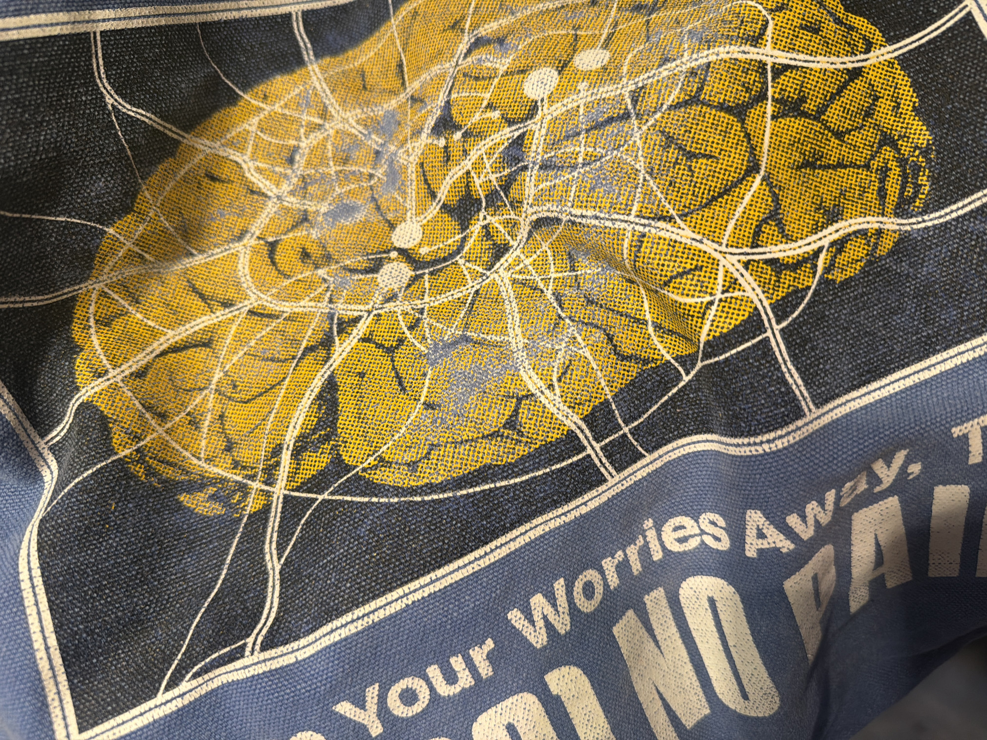 The image shows a close-up, angled view of a blue fabric, likely a tote bag or piece of clothing, with text and a graphic print. The text, in large white sans-serif font, reads "Your Worries Away" and partially shows "NO PAIN". The graphic is a yellow and dark grey abstract design resembling a network of interconnected lines and circular shapes, superimposed on a dark blue background. There is no discernible location in the city of Khimki, Russia, visible in this image. The fabric appears to be textured, with a visible weave. The lighting is somewhat uneven, creating shadows and highlights on the fabric.
