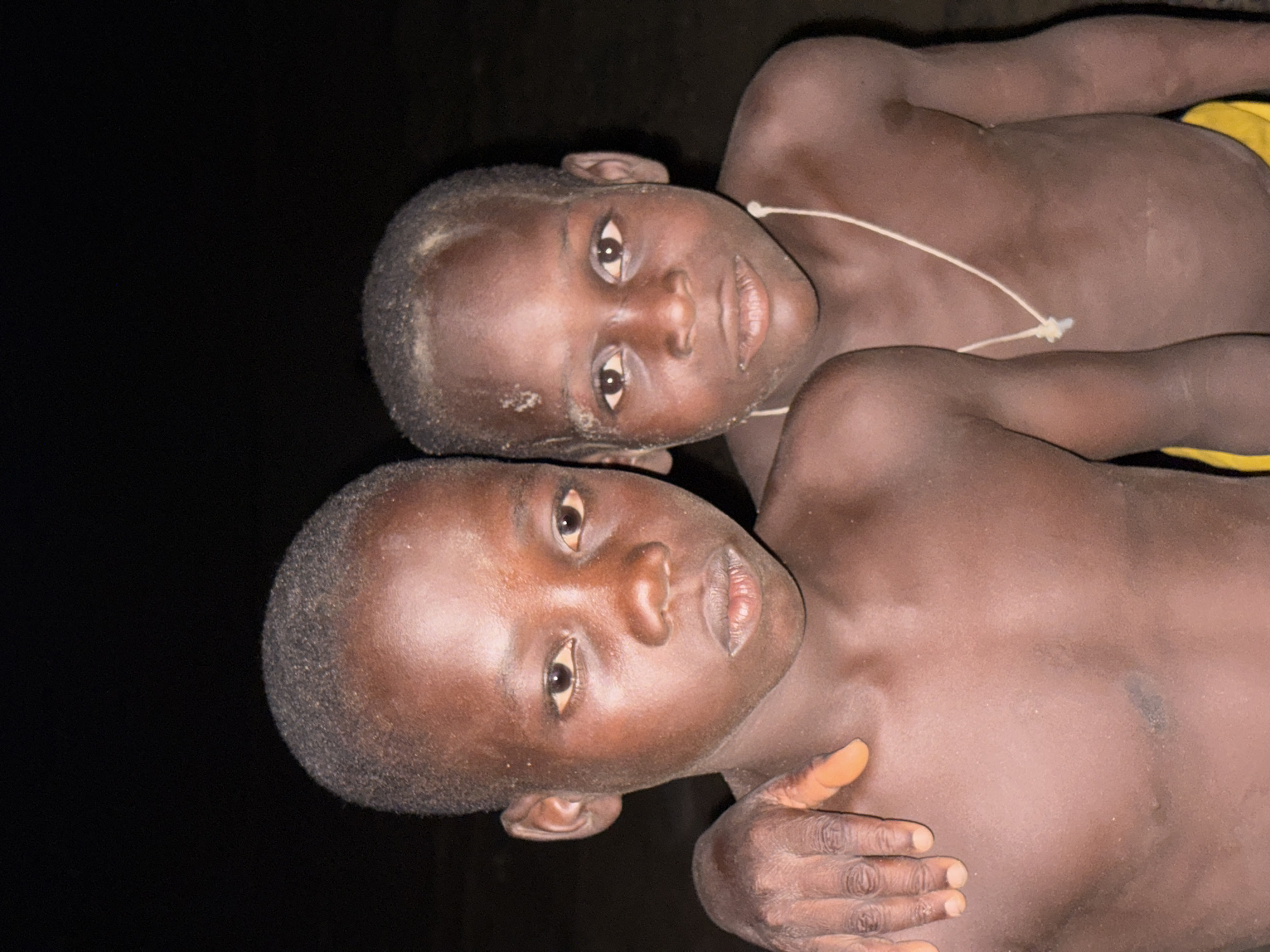 Two shirtless, dark-skinned boys are lying down close to each other. One boy is on his back, with his hand on the other boy’s torso. The other boy is on his side, facing the camera. A white cord is visible around the neck of the boy on his back. The background is black, but there is no additional information to connect the image to the city of Ikoyi, Nigeria.