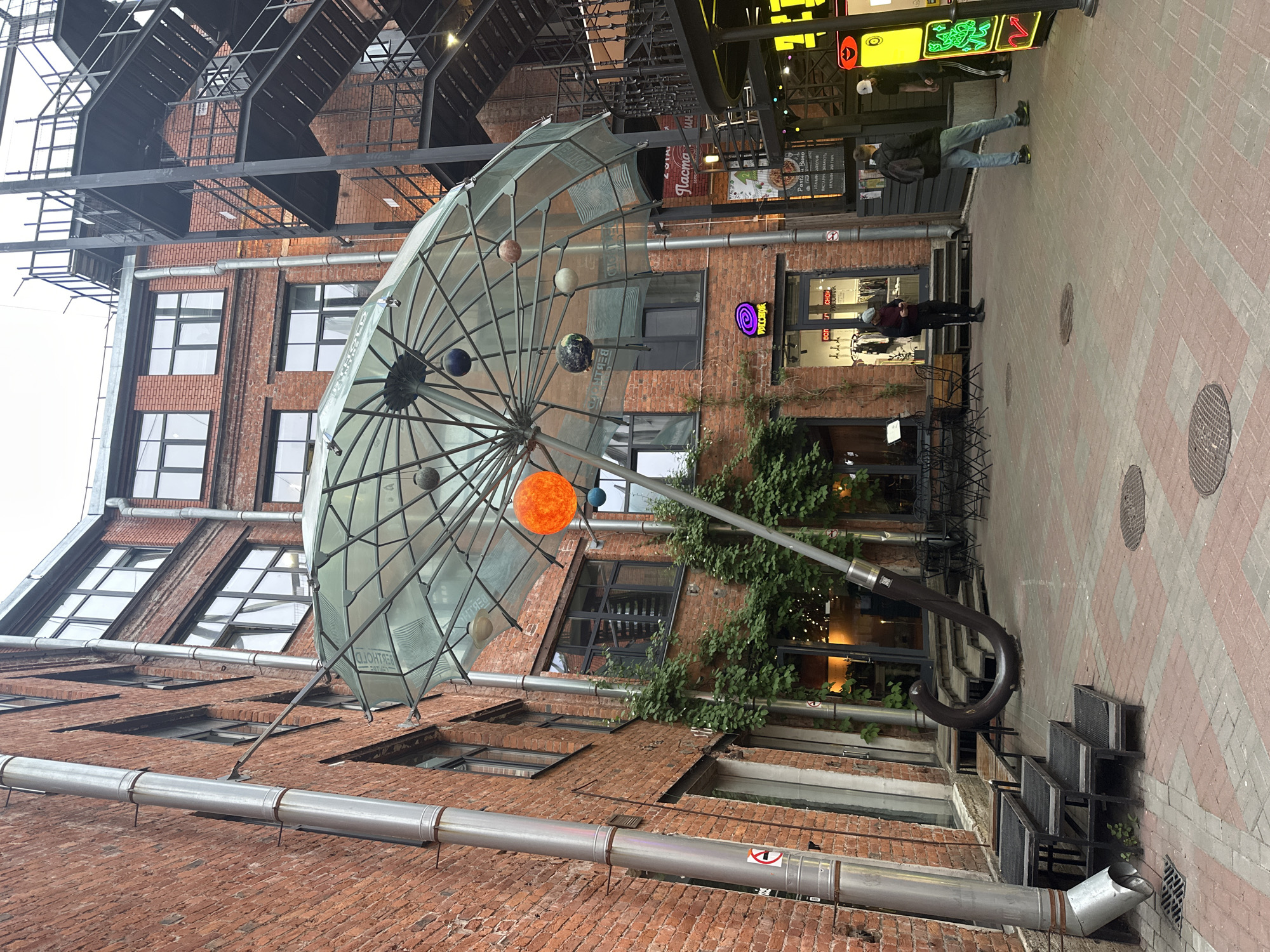 The image shows a narrow courtyard in Saint Petersburg, Russia. The courtyard is enclosed by brick buildings with multiple windows and external metal staircases. A large, artistic representation of an umbrella with models of planets attached is suspended in the center of the courtyard. The umbrella has a transparent canopy and a curved metal handle that reaches the ground. The ground is paved with red and grey bricks. Several metal chairs and tables are visible near an entrance to one of the buildings, suggesting a café or restaurant. People are present near the entrance, including one individual standing near the entrance and another passing by. There are also signs and lights indicating the presence of commercial establishments. A silver pipe runs along the base of one of the buildings.