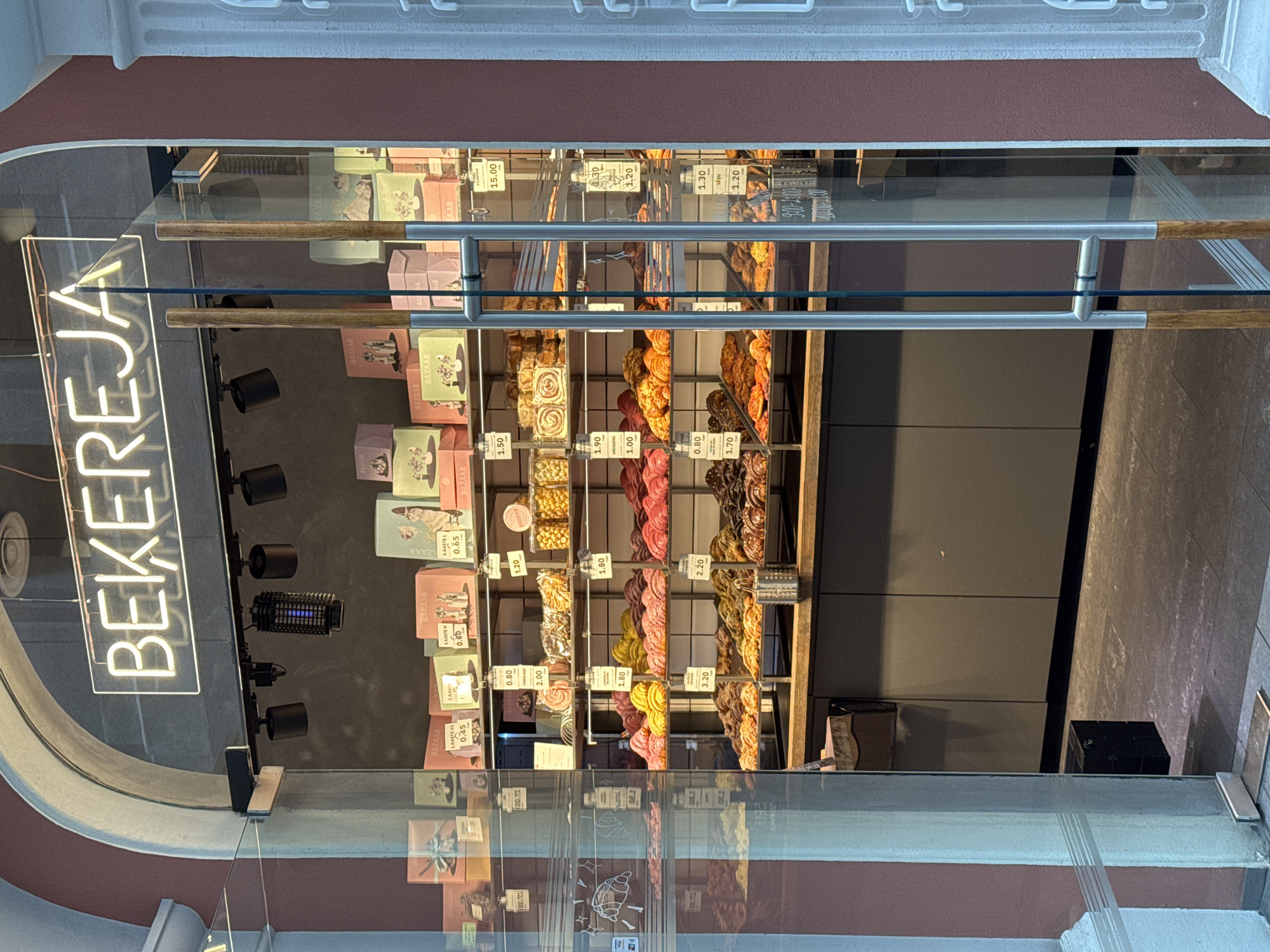 The image depicts a bakery storefront, likely in Riga, Latvia, as indicated by the name "BEĶEREJA" in a neon sign. The storefront is primarily pink and grey. There is a transparent glass display case that shows bakery goods, including pastries and confections, arranged on shelves with price tags. There are also small gift boxes with the word "BAZAAR" printed on them. The prices range from 0.45 to 15.00. There is also a bug zapper hanging above the display.