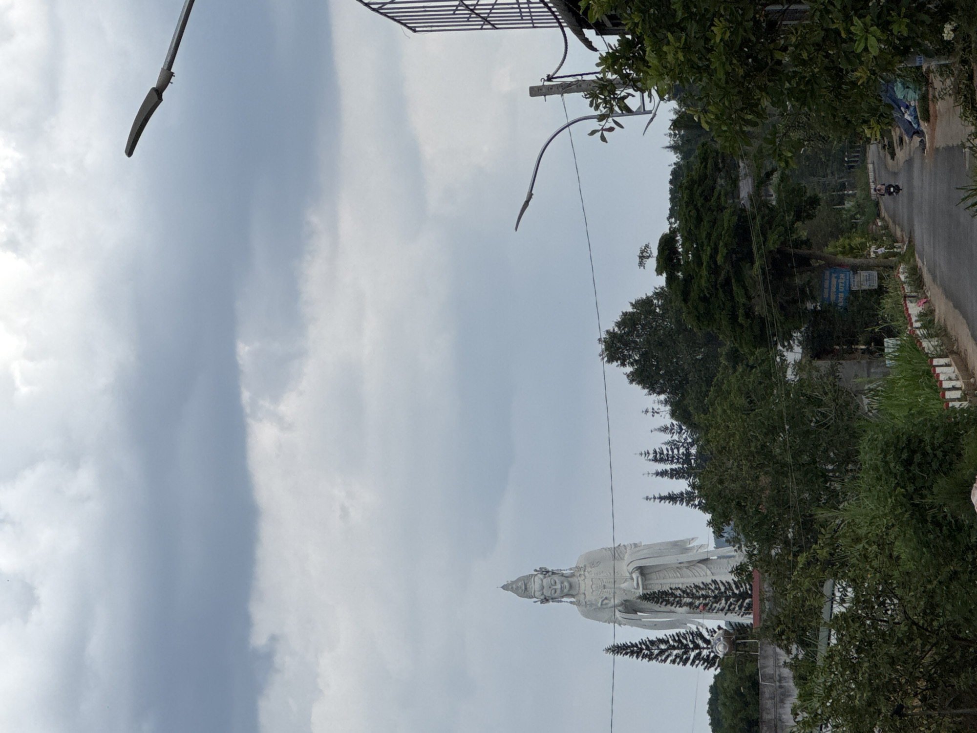 The image captures a partially obscured view of the Lady Buddha statue located in Nam Ban, Vietnam. The statue, a large white figure in a traditional robe and headdress, is positioned against a backdrop of a cloudy, overcast sky. The lower portion of the statue is framed by green trees and vegetation, suggesting it is situated in a park or garden setting. A road runs alongside the park, and a motorcycle can be seen driving on it. Street lamps are also visible.
