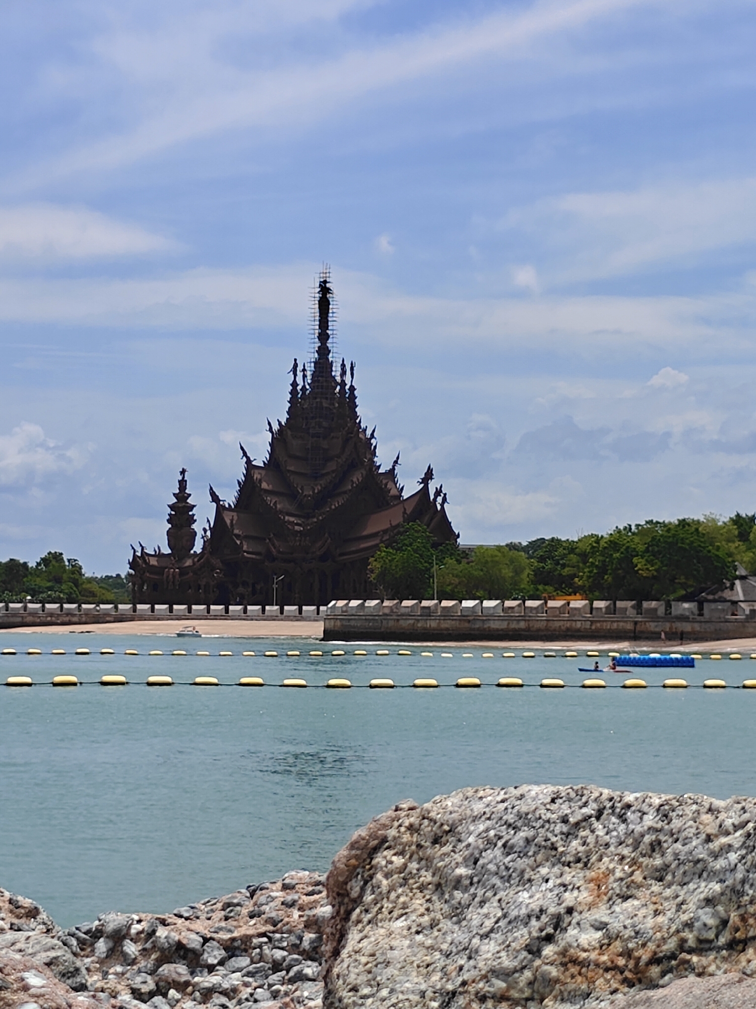 The image depicts the Sanctuary of Truth in Pattaya City, Thailand. The iconic wooden structure, characterized by its intricate carvings and tiered roofs, is seen against a backdrop of a cloudy blue sky. In the foreground, a calm body of water is visible, marked by a line of yellow buoys. A sandy beach and lush green trees are present behind the buoys, leading up to the Sanctuary. A small boat can be seen on the water near the beach, and a blue floating platform with two individuals on it is also present. The composition is completed by rough rocks in the immediate foreground, suggesting the viewer is positioned on a rocky shoreline.