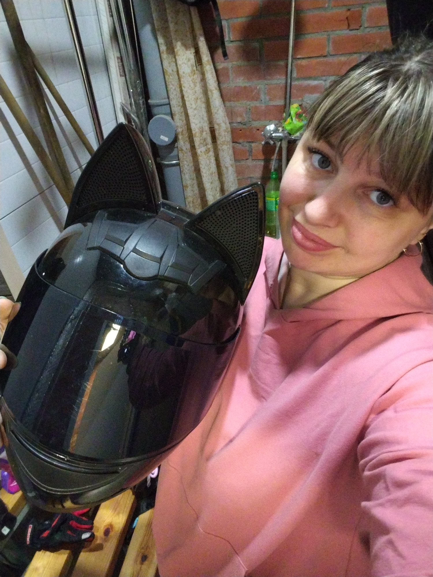 A woman takes a selfie indoors, likely in a storage or utility area, possibly in Rusanovo, Russia. She looks directly at the camera with a slight, playful smile, her lips lightly parted. The woman has light skin, blue eyes, and light brown hair with blonde highlights styled into bangs. She is wearing a light pink hooded sweatshirt and small gold hoop earrings.
She holds a glossy black motorcycle helmet featuring prominent black cat ears on its top and a dark visor. The helmet reflects some ambient light and indistinct shapes, including hints of purple and white.
The background shows an eclectic mix of elements: a reddish-brown brick wall to the right, adorned with a silver tap or faucet and some vertical pipes. To the left, a white tiled wall is visible, with several long, light-colored wooden poles leaning against it. A green bottle is partially visible near the woman's shoulder, behind the helmet. At the bottom left of the frame, a pair of red and black shoes rests on wooden slats, possibly part of a pallet. The indoor setting, with its varied wall textures and practical items, suggests a garage, workshop, or storage room. The lighting appears to be artificial. The woman's expression, combined with the distinctive helmet, conveys a cheerful and lighthearted mood. No discernible text is visible in the image.