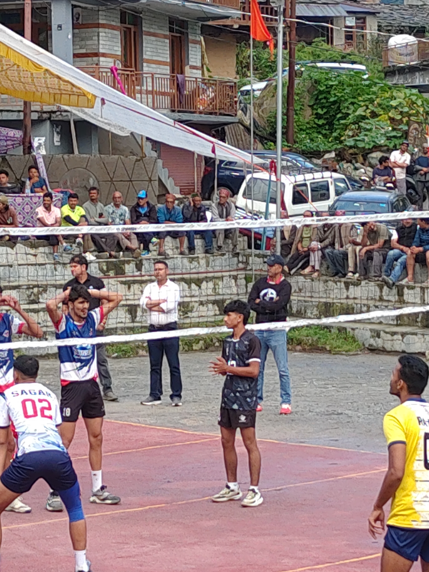 In Burwa, India, a volleyball game is in progress on an outdoor court. The court is marked with red and grey lines. A net divides the court, with players on both sides. In the foreground, three male athletes in athletic wear are on the court. One player on the left wears a white and blue jersey with the number '02' and blue shorts with knee pads. To his right, another player in a blue and white jersey with the number '10' on his shorts is in a defensive stance. On the right side of the net, a male player in a black and white patterned jersey and black shorts is looking upwards. Behind the players, spectators are seated on tiered concrete bleachers. The background shows buildings and lush green vegetation, typical of the mountainous region.