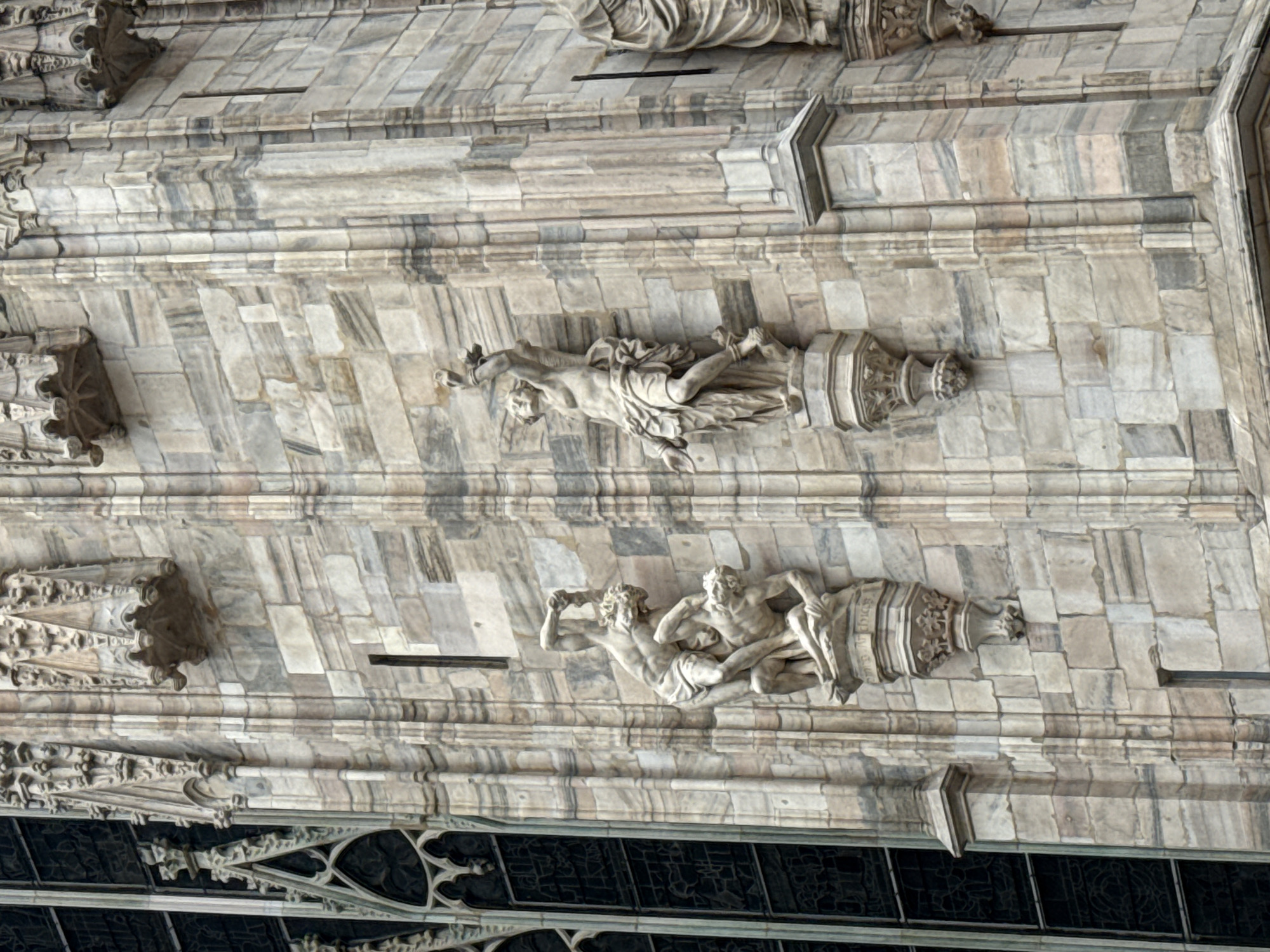 The image shows an exterior detail of the Duomo di Milano, a cathedral in Milan, Italy. The frame is angled, capturing a section of the façade with marble stonework. Two sets of sculpted figures are visible. One appears to depict a single figure, possibly bound or constrained. Below, another sculpture shows two figures engaged in what seems to be a struggle or combative interaction. Below the structures is the top of the window, decorated with ornate patterns, providing contrast to the aged stone.