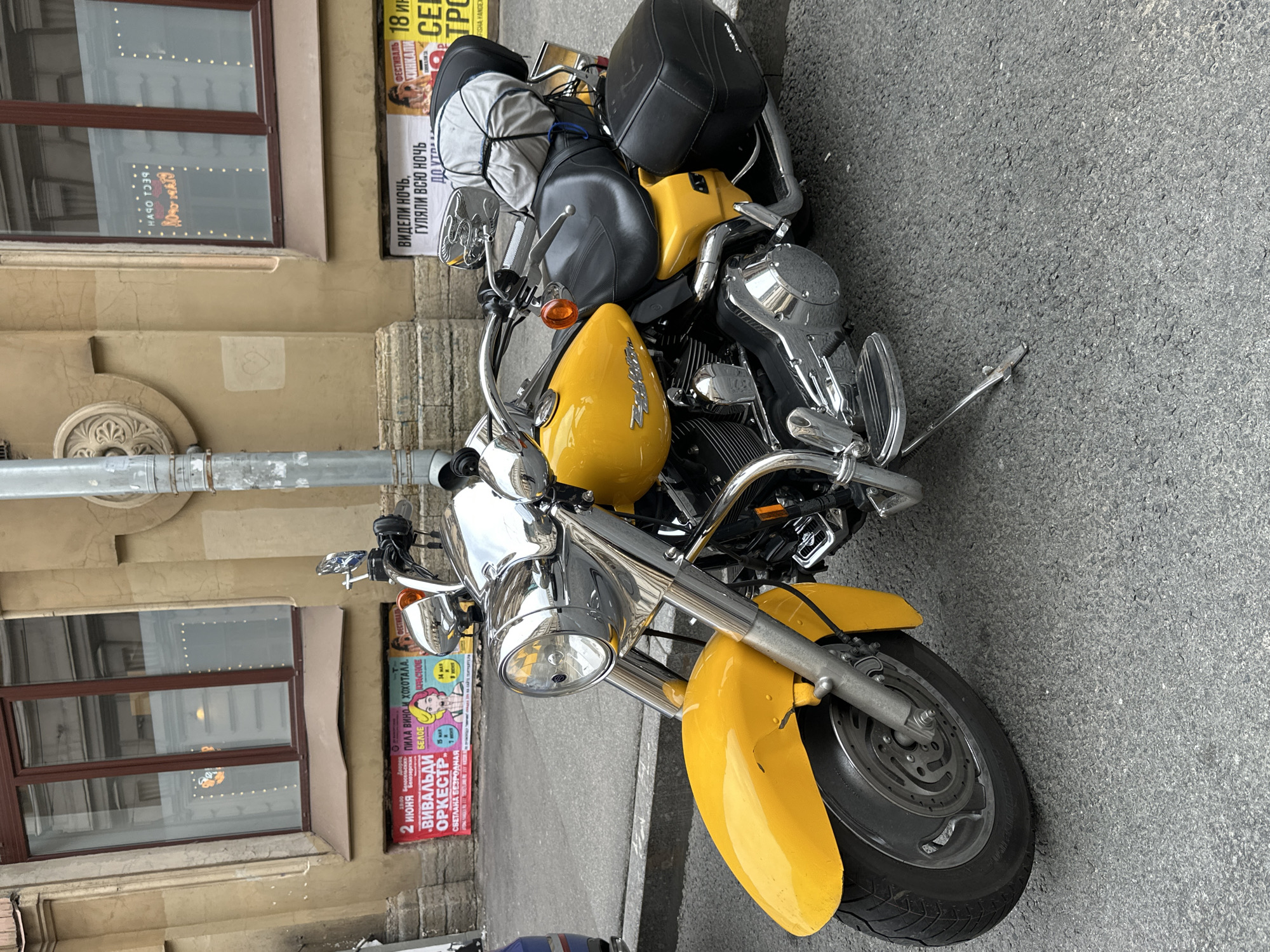 A yellow Harley Davidson motorcycle is parked on a city street next to a building with windows and advertisements. The motorcycle has luggage strapped to the back and a kickstand holding it upright. The building is light beige with brown window frames. Two advertisements are visible: one for the "Vivaldi Orchestra" on June 2nd and another with the phrases "ВИДЕЛИ НОЧЬ" and "ГУЛЯЛИ ВСЮ НОЧЬ" - which is Russian for "Saw the night" and "Walked all night". The scene appears to be in Saint Petersburg, Russia.