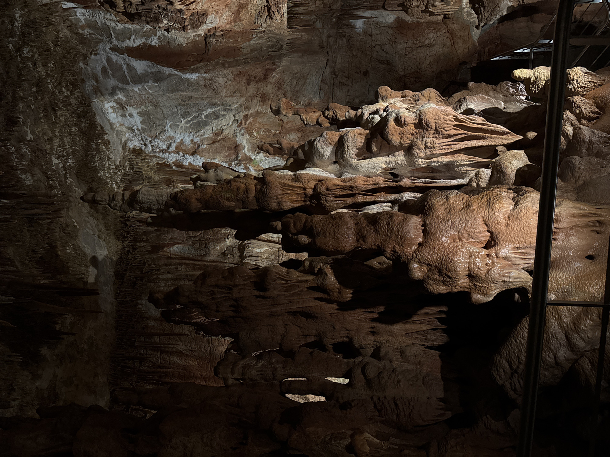 The image shows a section of the cave in Petralona, Greece. The cave walls are rough, textured, and layered, with variations in color from brown to white. The structures are rock formations, specifically stalactites and stalagmites. A metal railing is present on the right side of the image. The lighting in the cave appears dim, casting shadows and highlighting the natural contours of the formations.