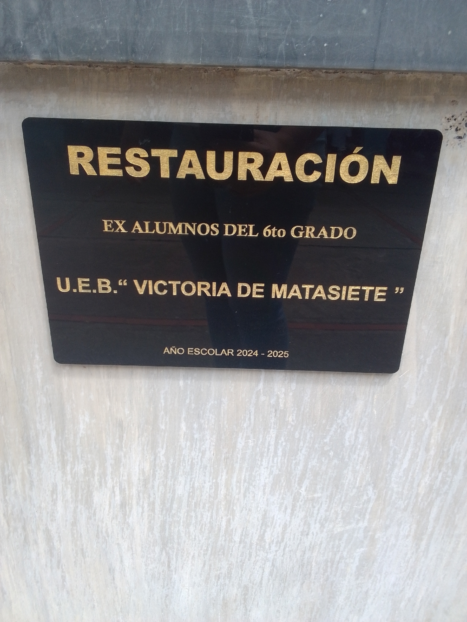 This media file contains a close-up, slightly angled shot of a black plaque mounted on a light-colored, textured wall. The plaque has gold lettering. The top line of text reads "RESTAURACIÓN". Below that, it says "EX ALUMNOS DEL 6to GRADO". The next line is "U.E.B." VICTORIA DE MATASIETE "". The bottom line indicates "AÑO ESCOLAR 2024 - 2025". The wall behind the plaque appears to be made of concrete or stucco with vertical streaks and some imperfections. The top edge of the image shows a darker, smoother surface, possibly a metal trim or a different wall material. The lighting suggests an outdoor setting, as there are some shadows and the overall illumination is even. There are no people or distinct weather conditions visible. The scene appears to be a commemorative plaque in a public or institutional setting, likely related to a school or alumni association. The text indicates a restoration project by former students of the 6th grade at the "Victoria de Matasiete" educational unit during the 2024-2025 school year.