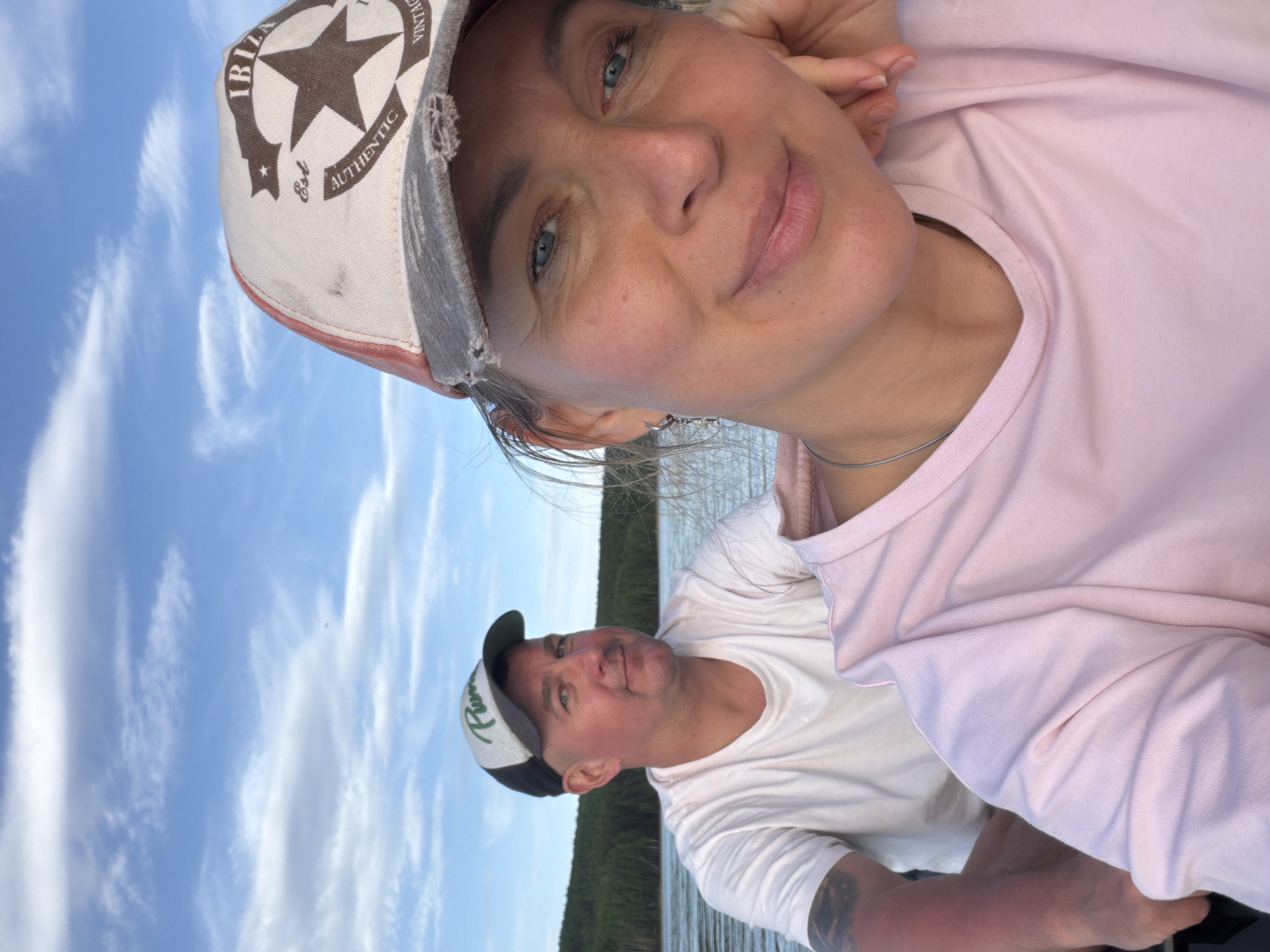 A selfie shows a woman and a man on a boat. The woman is in the foreground, wearing a light pink shirt and a distressed baseball cap with a star design. The man is behind her, wearing a white t-shirt and a black and white baseball cap. The background features a body of water, a treeline, and a blue sky with clouds. The exact location in Chaykovsky, Russia, is not discernible from the image.
