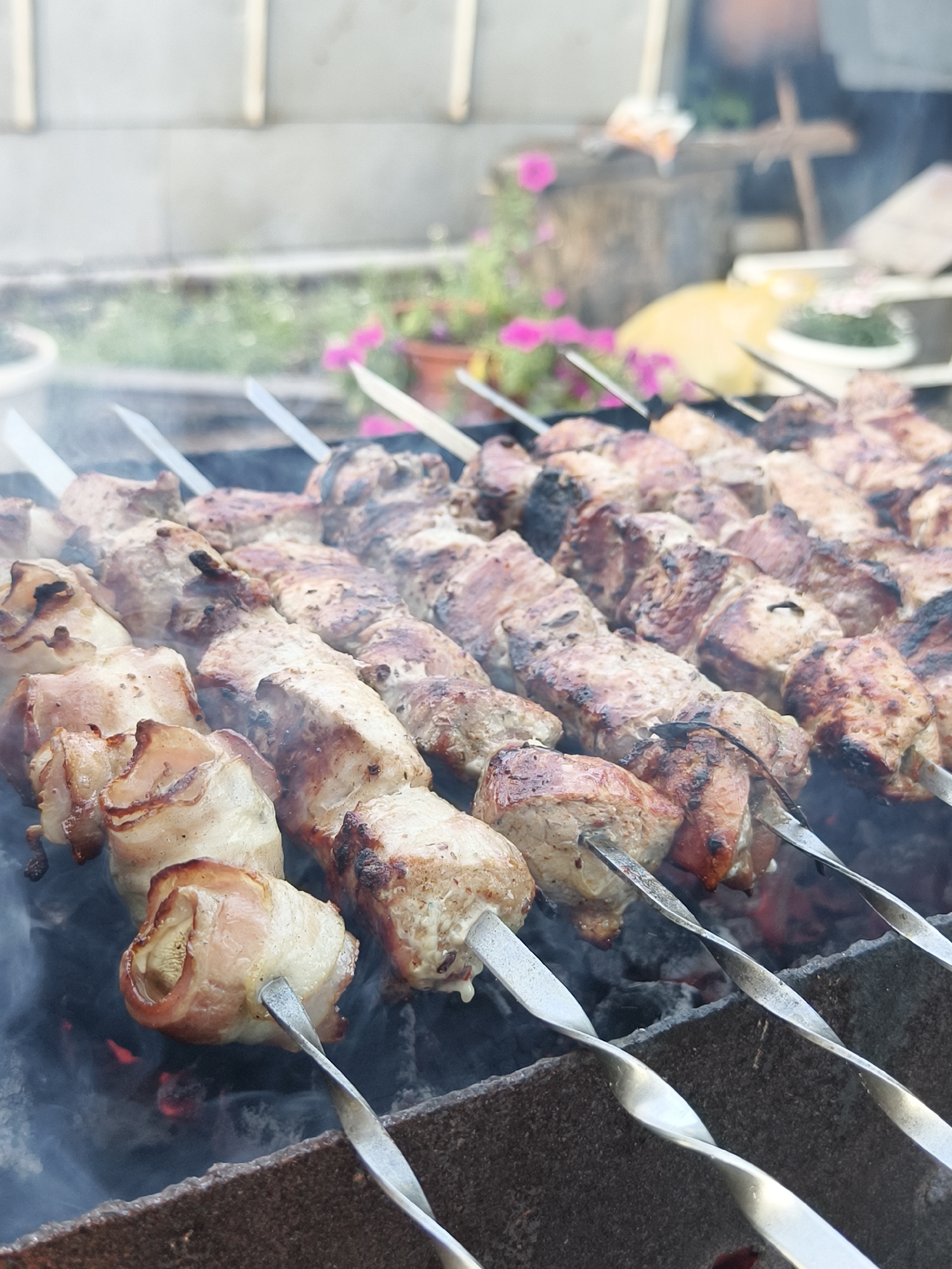 In Семилетка, Russia, skewers of meat are being grilled over hot coals. The meat, some of which is wrapped in bacon, is seasoned and appears to be pork. The skewers are arranged in rows and are actively cooking, with smoke rising from the grill. The background shows a blurred outdoor setting with some greenery and structures, suggesting a backyard or a casual outdoor gathering. The focus is on the food preparation, highlighting the process of cooking shashlik.