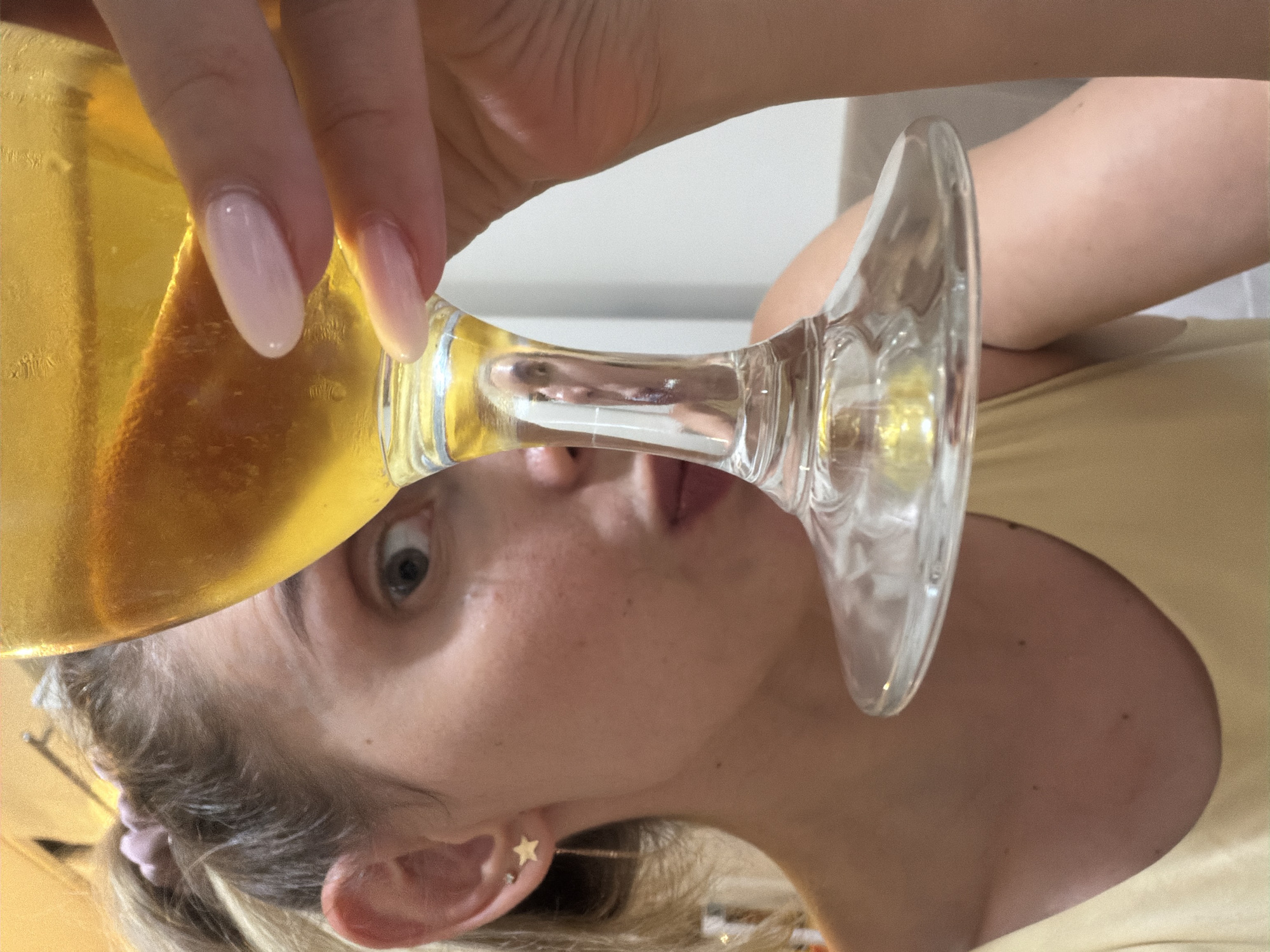 The image shows a close-up, vertically oriented shot of a woman holding a champagne flute filled with a golden-hued beverage. Her hand, with meticulously manicured light pink nails, grips the stem of the glass. The woman's face is partially obscured by the glass, but her eyes are wide and looking towards the camera, suggesting a moment of surprise or candidness. She is wearing a light-colored top and has blonde hair pulled back, with a small star-shaped earring visible on her ear. The background is softly blurred, with hints of a light-colored wall and a yellowish surface, possibly indicating an indoor setting. The lighting appears to be natural and bright. The scene conveys a sense of celebration or enjoyment, possibly during a social gathering, given the champagne flute. There is no visible text in the image. The location context provided is Budva, Montenegro, which doesn't offer specific visual cues within the image itself.