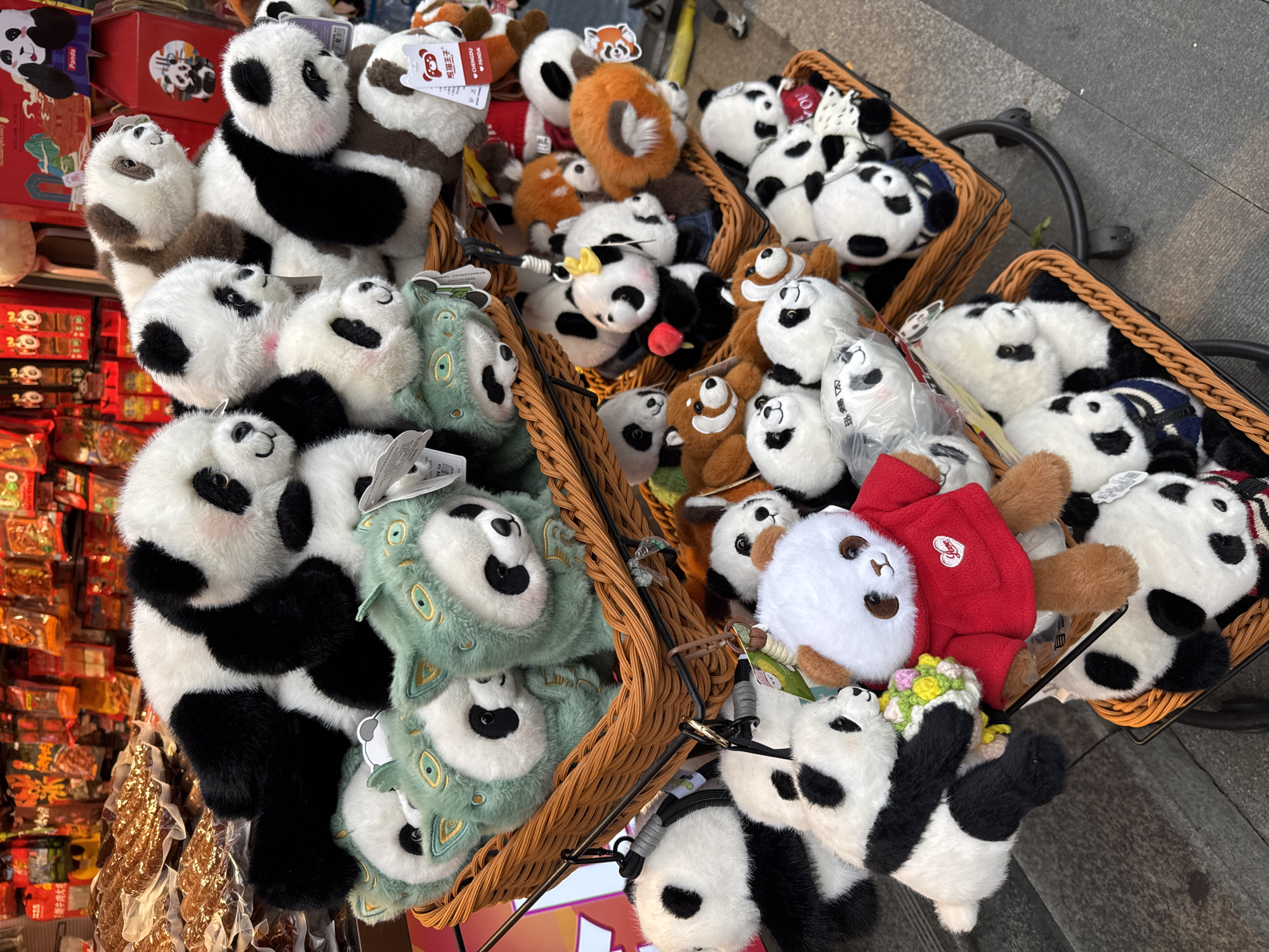 This image shows a collection of stuffed pandas and other animal plush toys displayed in woven baskets, likely for sale. The scene appears to be in a retail setting, with some food items visible in the background, suggesting a tourist area. The presence of tags with Chinese characters and the word "Chengdu" indicates that the location is likely in or related to the Jinjiang District, China, known for its panda-related tourism and merchandise. The objects primarily feature panda-themed plush toys in various designs, along with some non-panda animal plush toys. The baskets are placed on a cart or stand, suggesting a temporary or mobile sales setup. The overall environment implies a bustling, commercial area catering to tourists interested in local souvenirs and panda-related items.