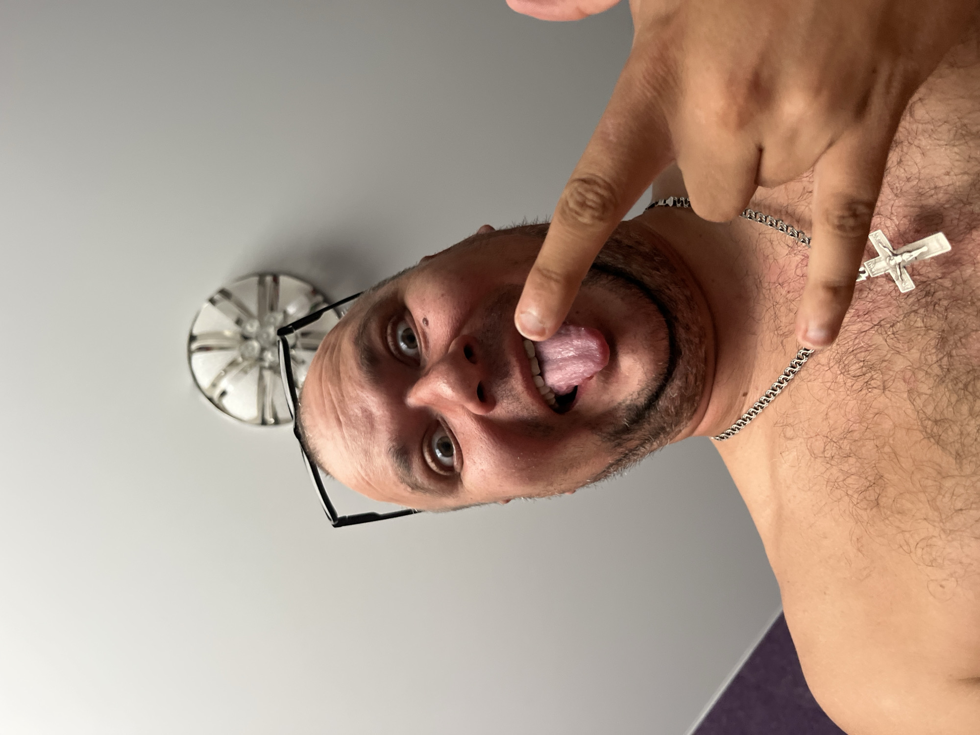 The image is an upside-down, close-up, eye-level shot of a man. He is wearing a silver chain with a cross pendant. His tongue is sticking out, and his eyes are wide open. He is wearing black-rimmed glasses. His skin appears fair, and he has dark hair and a beard. A light fixture with a decorative pattern is visible in the upper left corner. The background is a plain, light-colored wall. The lighting suggests it could be daytime indoors. The man's expression is playful or mischievous. There is no visible text in the image.