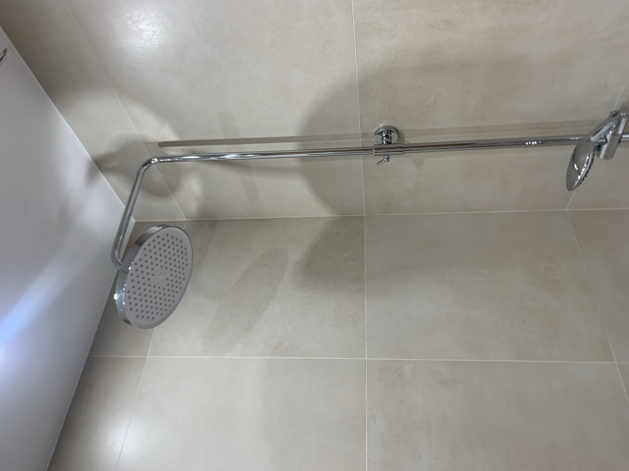 The image shows a close-up, low-angle view of a modern chrome shower fixture mounted on a tiled wall. The showerhead is a circular, flat design with numerous small spray nozzles, attached to a curved metal arm extending from a vertical pole. A second, smaller handheld showerhead is mounted lower on the pole. The wall is covered in large, square, light beige tiles with thin, lighter grout lines. The ceiling is visible at the top, illuminated by a light source that casts a subtle blue and white glow. The scene is clean and appears to be in a bathroom setting. There are no people or any indication of actions or interactions. The location is not specified in the image itself, and therefore cannot be determined to be Vladivostok, Russia.